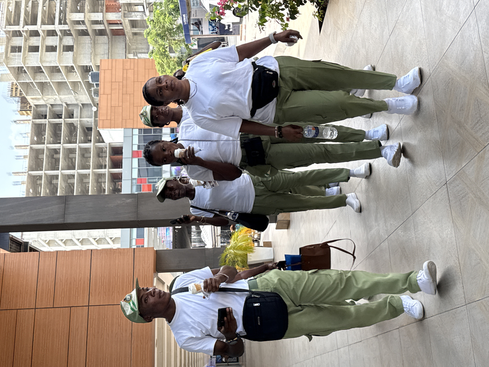 The image shows a group of five people standing in a line on a tiled walkway. All are wearing white T-shirts, olive-green pants, and white sneakers. Each person is carrying a black shoulder bag. The person in the front is holding a cell phone in one hand and what appears to be an ice cream cone in the other. He is also wearing a green and white hat. The second person is holding an ice cream cone in one hand. The third person is holding a water bottle in one hand. The fourth person is holding a white object. The fifth person has one arm up. A large construction building is visible in the background. A building with colorful windows is visible in the midground. The location is Alaguntan, Nigeria.