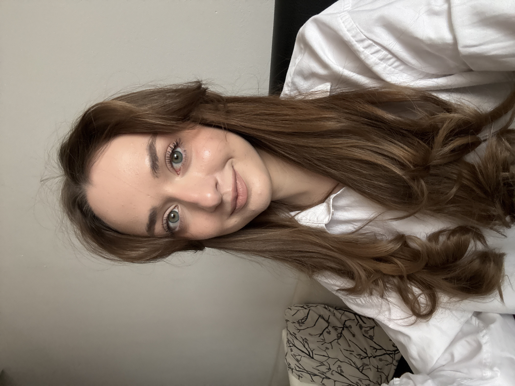 The original image, displayed rotated 90 degrees counter-clockwise, presents a close-up, selfie-style portrait of a young woman in an indoor setting, contextually identified as Kragujevac, Serbia.
**Subjects:** The primary subject is a young woman.
**Objects:** She is wearing a white collared shirt. Her long, wavy, medium-brown hair is prominent. In the background, there's a plain light-colored wall, a dark surface (possibly a headboard or furniture), and a patterned fabric, likely a pillow or bedding with a design of dark branches on a lighter background.
**Scene/Setting:** The scene is an interior space, most likely a bedroom or a comfortable indoor area.
**Activity or event taking place:** The woman is posing for a close-up photograph, possibly a selfie.
**Notable details:**
* **Time of day:** Indeterminate, as the lighting is soft and even, typical of indoor artificial light or diffuse daylight.
* **Weather:** Not discernible from the image.
* **Location cues:** No specific visual cues within the image indicate Kragujevac, Serbia, but this is the provided context.
* **Emotions:** The woman displays a subtle, closed-mouth smile and looks directly at the camera, conveying a gentle and pleasant emotion. Her light blue-green eyes are open and engaging.
**Visible text:** There is no visible text in the media file.
