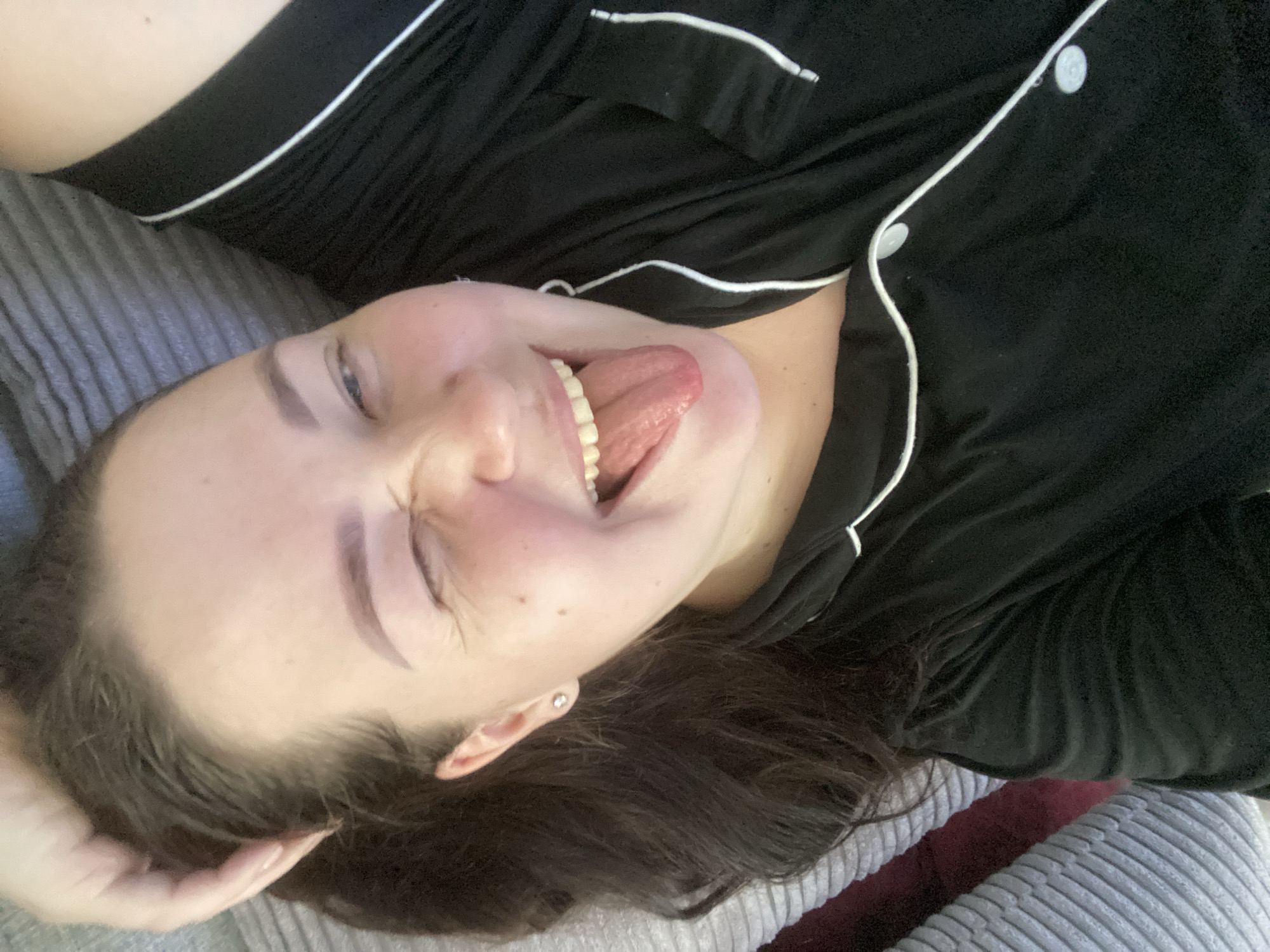 A woman is lying on a gray striped pillow. She is wearing a black pajama top with white piping and white buttons. Her head is tilted back, her eyes are closed, and her tongue is sticking out. She has dark hair and light skin. Her right hand is touching her hair. The background is indistinct. The location is unknown.