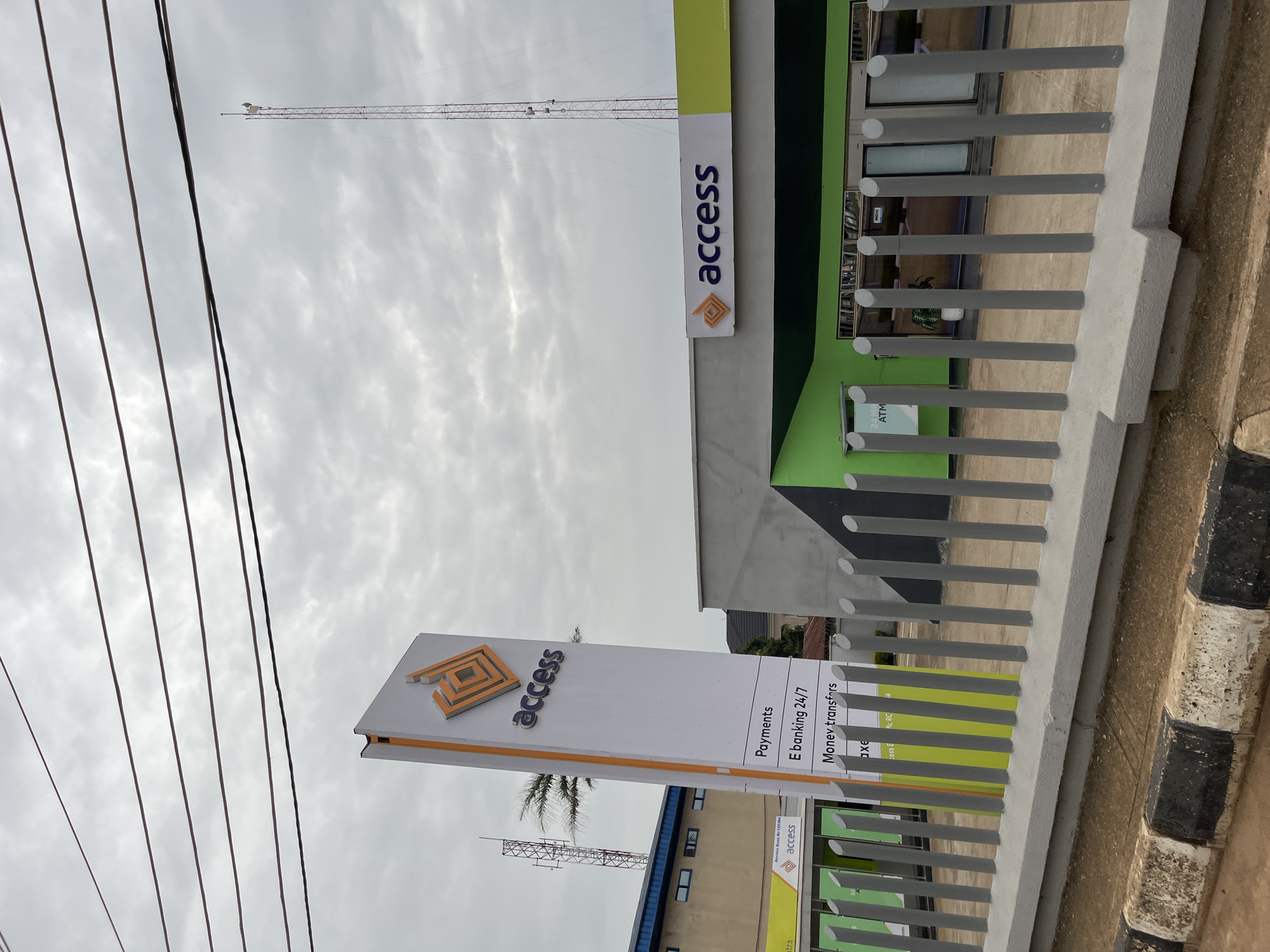 This is a ground-level, eye-level, slightly angled exterior shot of an Access Bank branch in Bukuru, Nigeria, under an overcast sky. A tall, white rectangular sign with an orange geometric logo and the word "access" in silver lettering stands to the left of the bank building. Below the logo, a list of services is visible: "Payments," "E banking 24/7," and "Money transfers." The main bank building features a gray facade with a prominent green section on the right, where another "access" logo is displayed. The entrance to the bank has glass doors and windows. In front of the bank is a gray metal fence. The foreground shows a curb with black and white markings. Power lines stretch across the top of the frame, and a tall communication tower is visible in the background on the right. Another smaller tower and part of a building are on the left. The environment appears to be a street in a city. There are no people visible in the image.