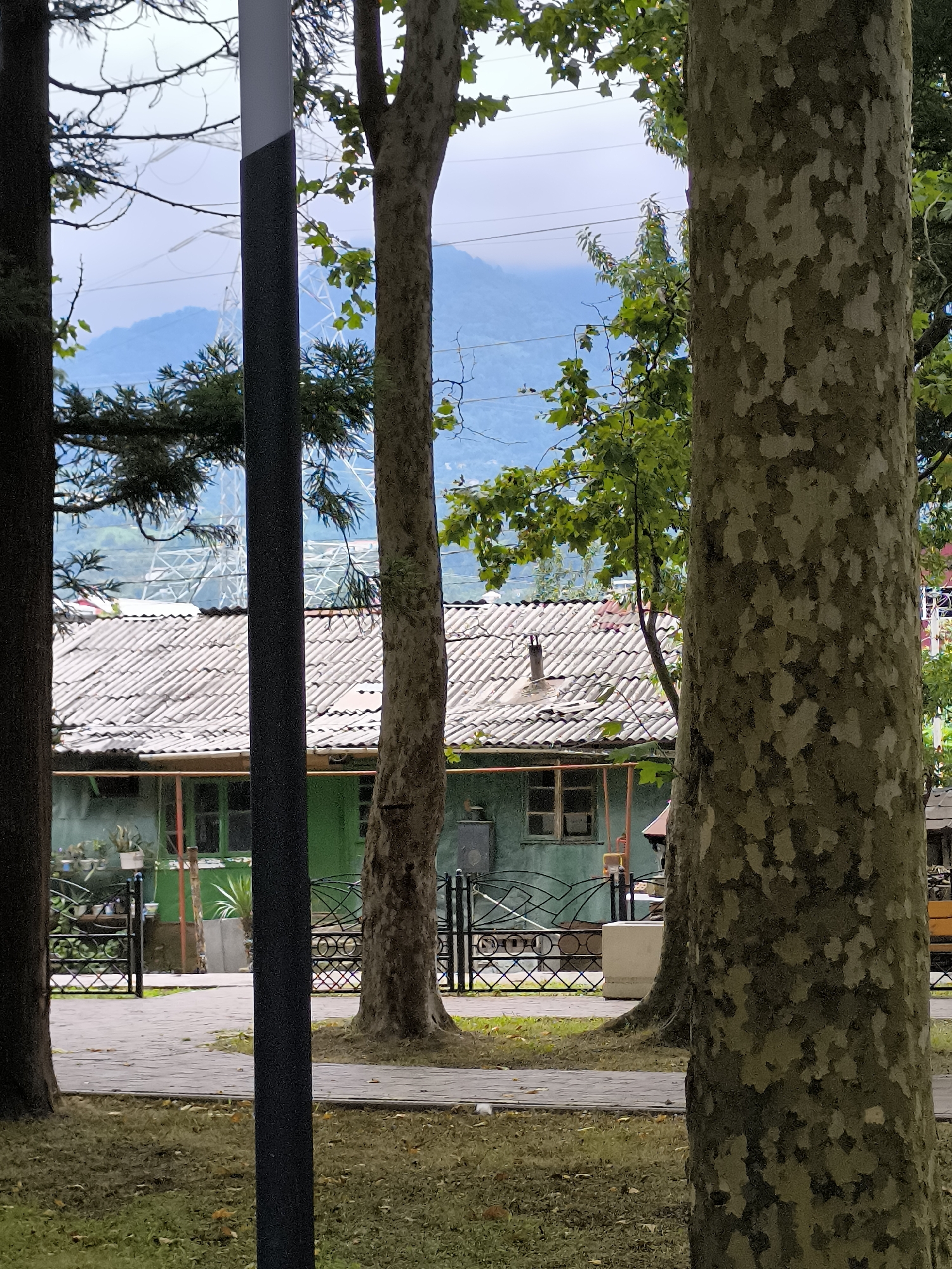 The image depicts a park scene with lush green trees in the foreground and a glimpse of a residential building in the background, set against a backdrop of rolling hills and a cloudy sky. In the mid-ground, a single-story building with a grey tiled roof and green walls is visible. A black metal fence with an intricate design separates the building from a paved pathway. The overall ambiance is serene and natural. The image is taken in Batumi, Georgia.