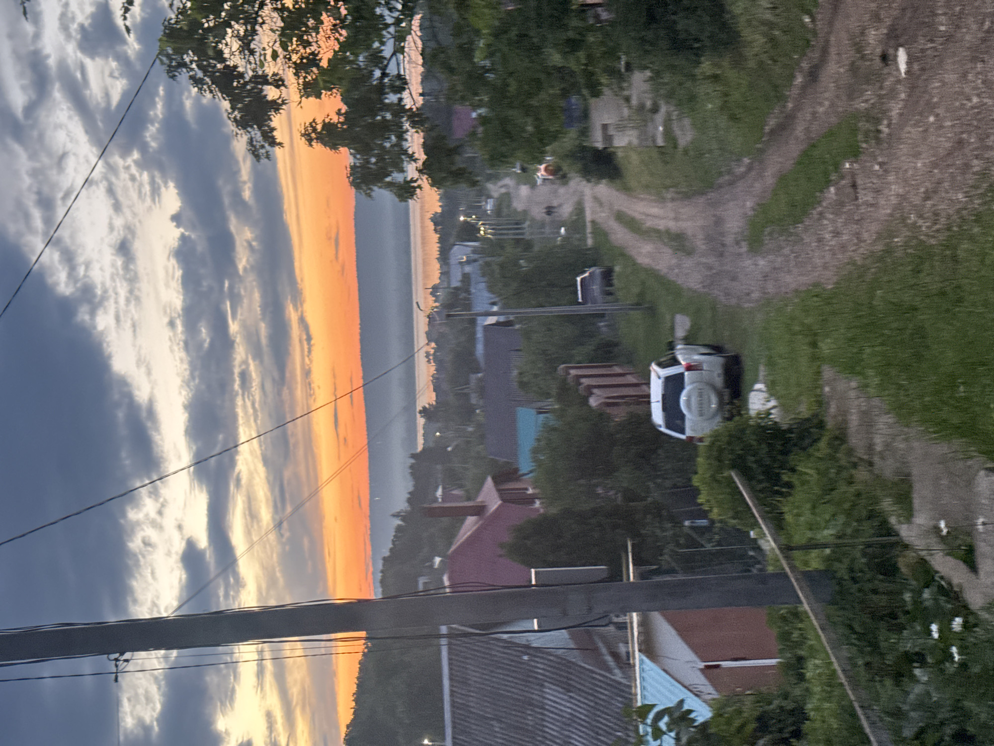 The image depicts a rural village scene at dusk. A dirt road curves through the foreground and middle ground, lined by grass and vegetation. Several houses with various roof styles are visible on either side of the road. A white SUV is parked on the left side of the road. Power lines crisscross the sky, and a utility pole stands prominently on the left. In the background, a body of water stretches towards the horizon, with a hazy outline of trees and buildings along its shores. The sky is filled with dramatic clouds, tinged with orange and yellow from the setting sun. The overall impression is of a quiet, pastoral evening in a remote settlement.