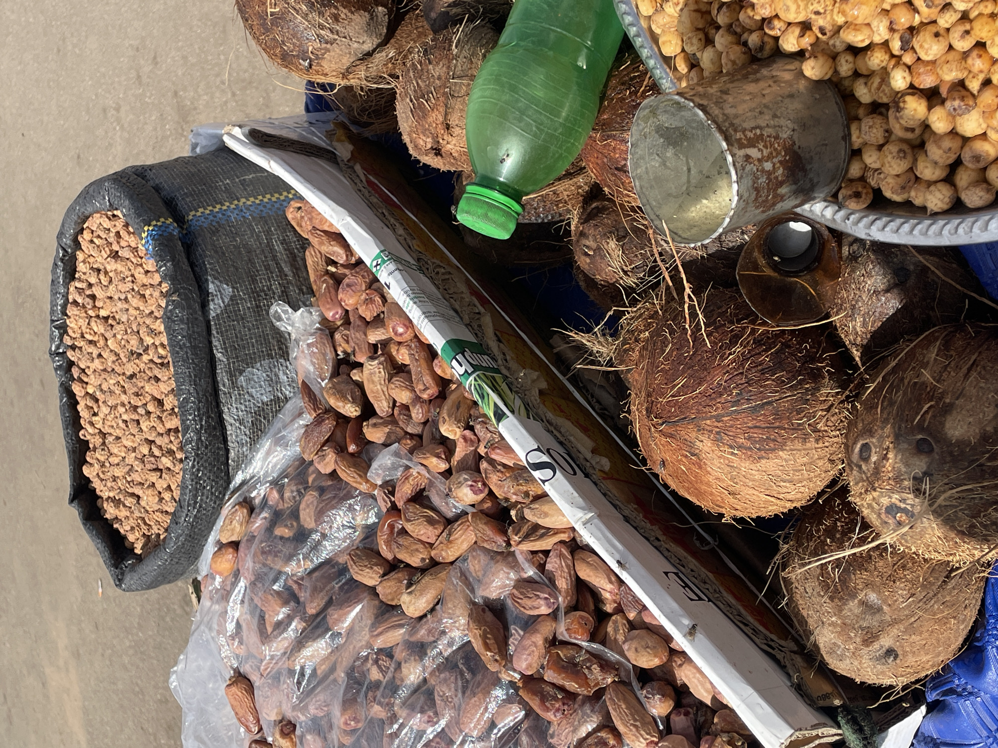 The image shows a high-angle view of a street vendor's cart in Bukuru, Nigeria. The cart displays a variety of items for sale, including:
* Several whole coconuts.
* A bag of small, light-brown, spherical nuts.
* A large plastic bag filled with dates.
* A green plastic bottle.
* A metal measuring cup.
* A tray of small, round snacks or nuts.
* A newspaper.
The items are arranged in a pile on a blue tarp covering the cart. The cart is parked on a paved surface.