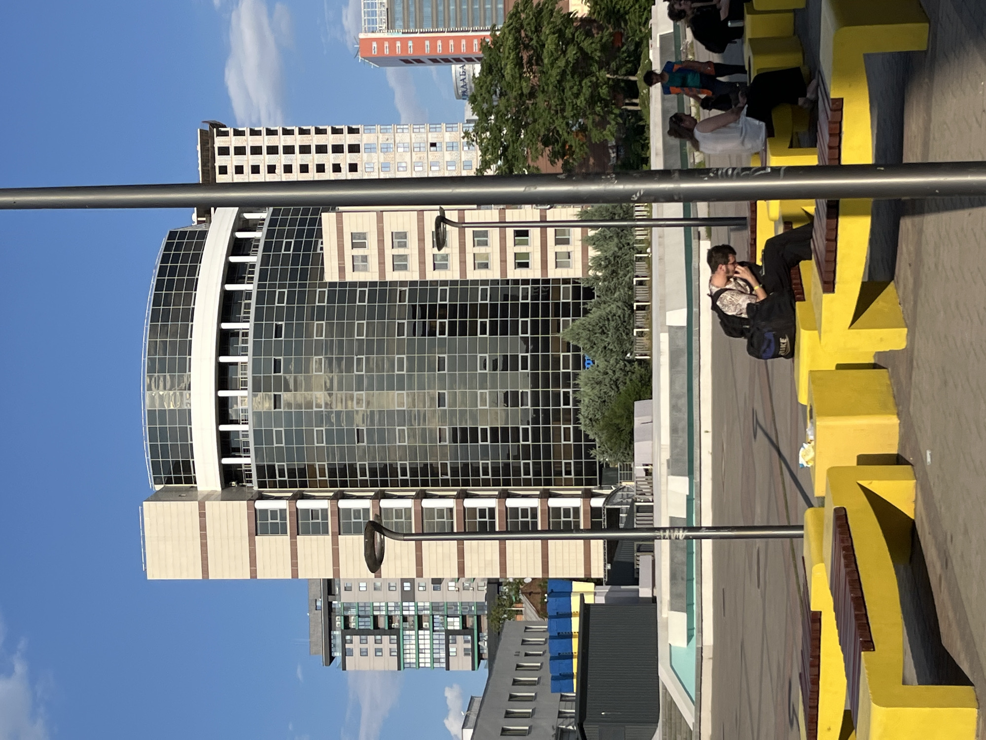 The image depicts a street scene in Dnipro, Ukraine, featuring modern architecture and a few individuals. In the foreground, a series of bright yellow benches are arranged along a paved area. Two people are seated on one of these benches, appearing relaxed. One person, wearing dark trousers and a patterned shirt, is looking down at their phone. The other person, in a white top and sunglasses, is looking to the side.
Behind the benches, a large, curved building dominates the mid-ground. This structure is characterized by its extensive glass facade, reflecting the clear blue sky. The building's design is modern and sleek, with horizontal white accents breaking up the glass panels.
Further in the background, several other buildings are visible. One is a tall, slender red and white structure with many windows. Another is a more utilitarian, multi-story building with a grid-like pattern of windows. The scene is bathed in natural light, suggesting it is daytime, likely late afternoon given the shadows. The sky is mostly clear with a few wispy clouds. The overall impression is one of a contemporary urban environment. There is no discernible text visible in the image.