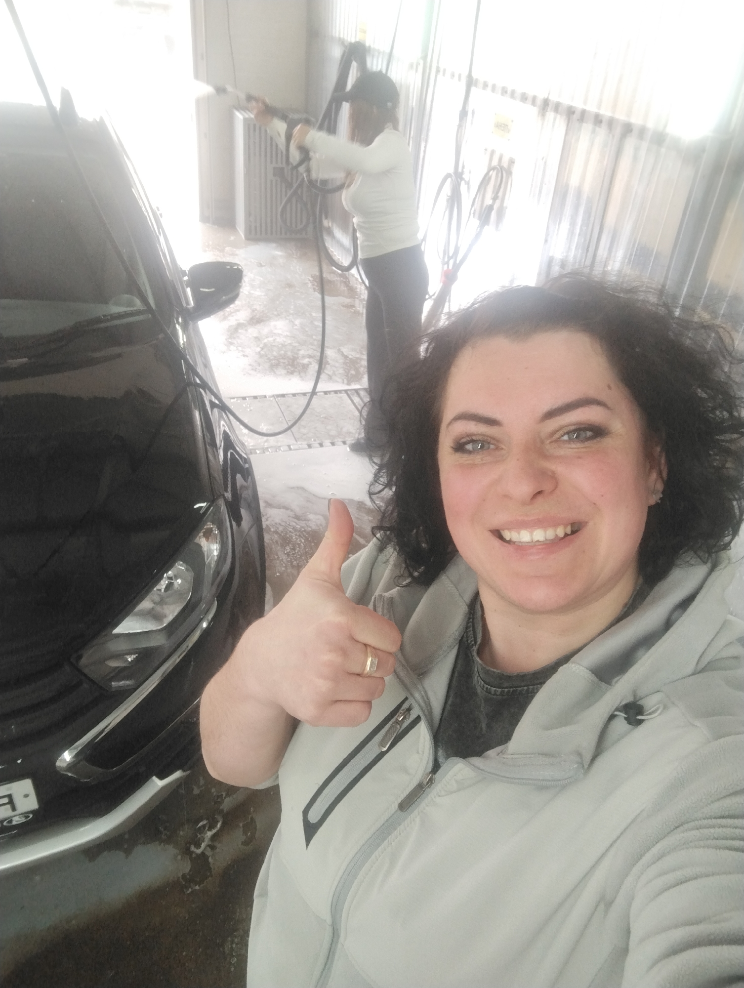 The image shows a woman taking a selfie at a self-service car wash in Votkinsk, Russia. The woman is fair-skinned with dark, curly hair and is wearing a light grey hooded jacket over a dark shirt. She is smiling and giving a thumbs-up. A black car is partially visible to the left, with water and soap suds visible on the ground. In the background, another person is using a high-pressure hose to wash a vehicle. The car wash bay has corrugated metal walls and bright lighting.