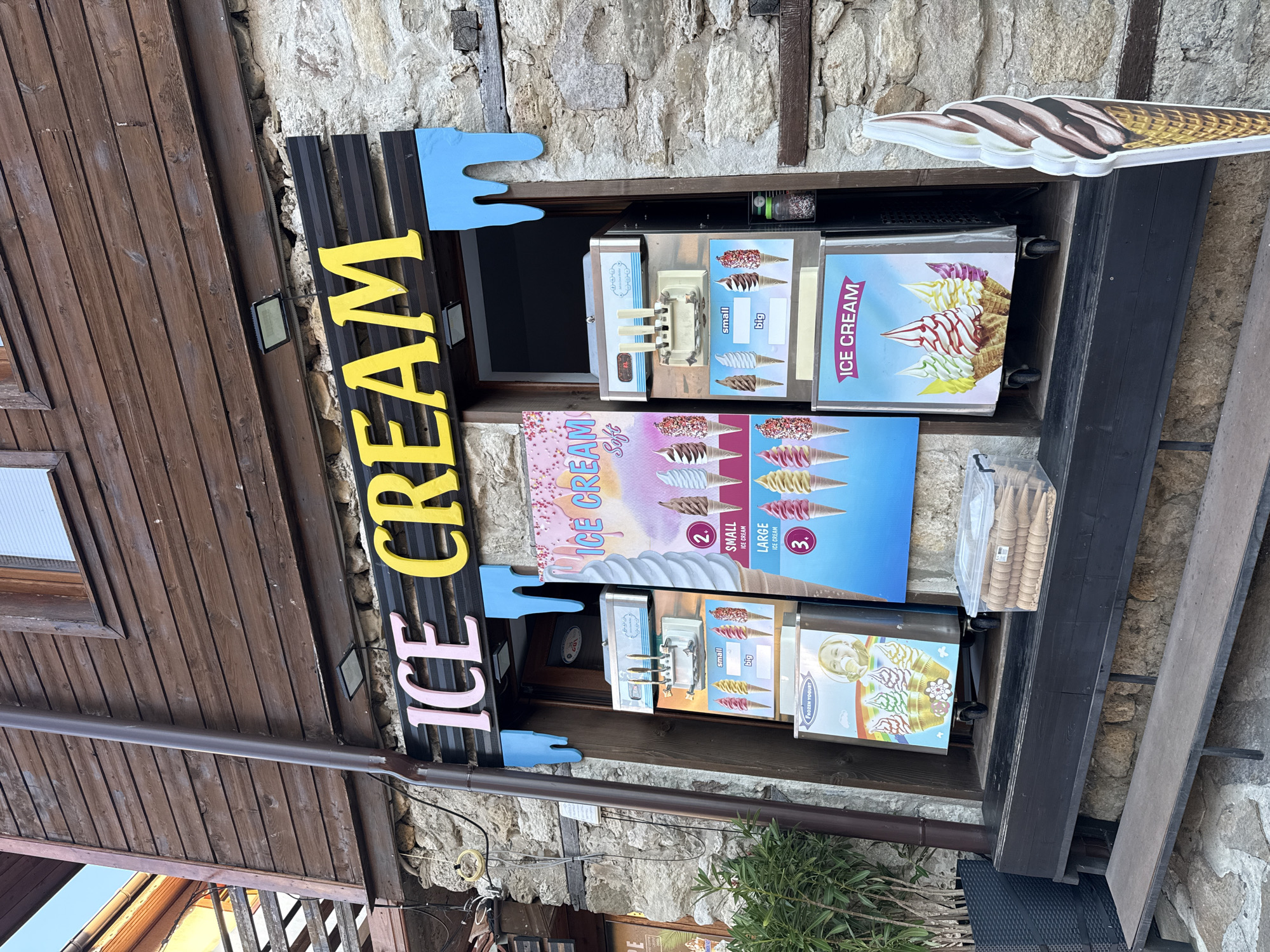 A facade of an ice cream shop in Nesebar, Bulgaria, is captured from a slightly low angle. The shop's name, "ICE CREAM," is prominently displayed in large, colorful letters, pink and yellow, against a black background. Blue, dripping decorations flank the signage. The building's exterior features a lower section constructed of rough, light-colored stone and an upper section clad in dark wooden planks.
In front of the shop, two ice cream machines are visible. The machine on the left has a poster advertising "Frozen Yogurt" with images of colorful soft-serve cones. The machine on the right displays various ice cream cone options, labeled "small" and "big." To the right of the machines, a large, standing cutout of a chocolate and vanilla swirl ice cream cone is positioned near the wall.
A dark wooden bench and a lower wooden step sit in front of the shop, with a clear plastic container filled with ice cream cones placed on the bench. A lush green potted plant is visible to the far left. The overall scene depicts a typical outdoor setting for an ice cream vendor in a historic town.
