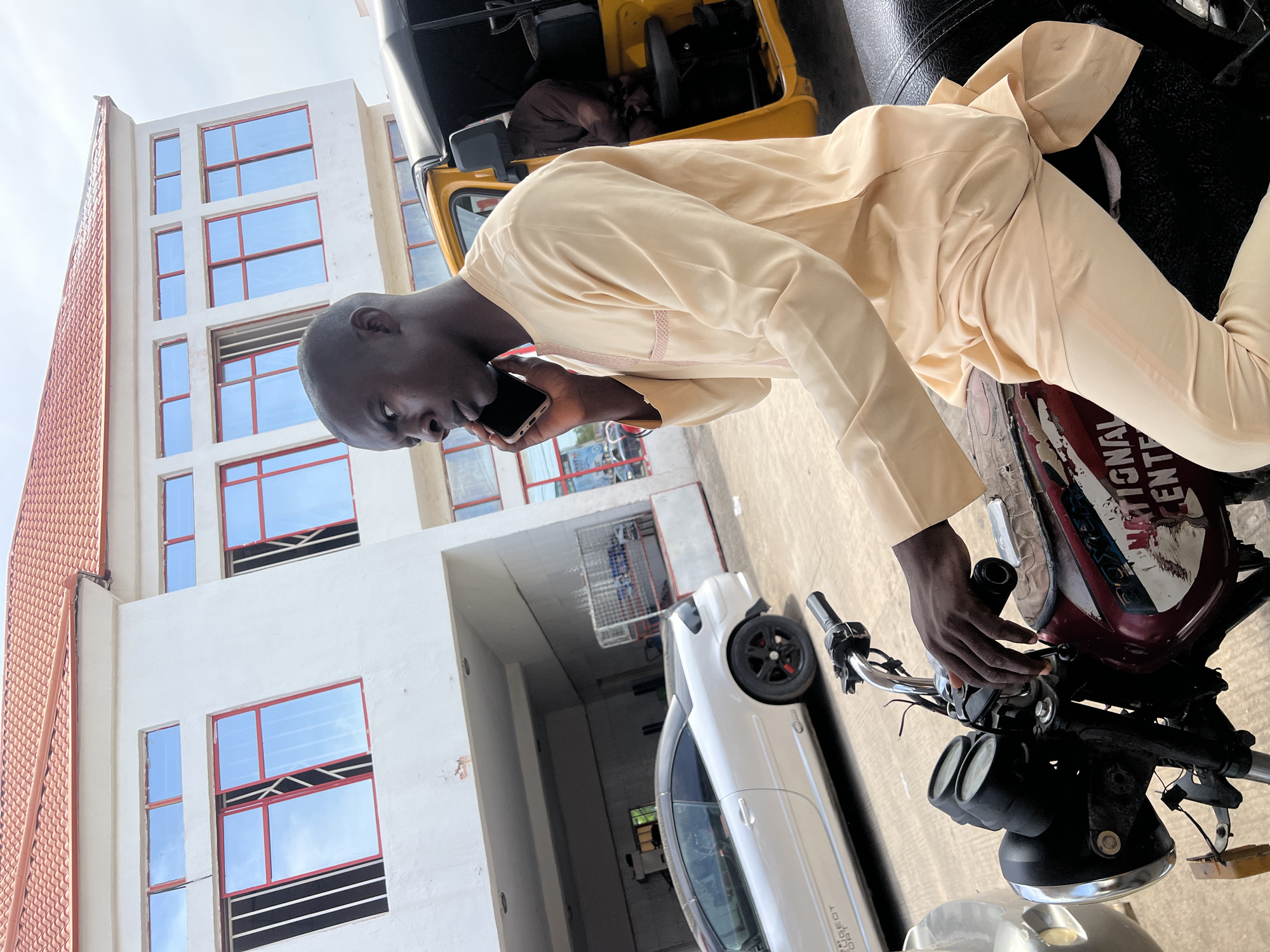 A person in a light-colored robe is talking on a smartphone while leaning on a motorcycle. The person's head is turned to the left, and their right hand is holding the phone to their ear. The motorcycle has a maroon and white fuel tank with some peeling stickers. The scene appears to be outdoors, with a white building featuring several windows with red frames to the left of the frame. A white car is parked next to the motorcycle. The sky is visible in the background, suggesting it is daytime. The overall impression is one of a casual, everyday moment. The context of Jalingo, Nigeria, suggests a potentially warm climate.