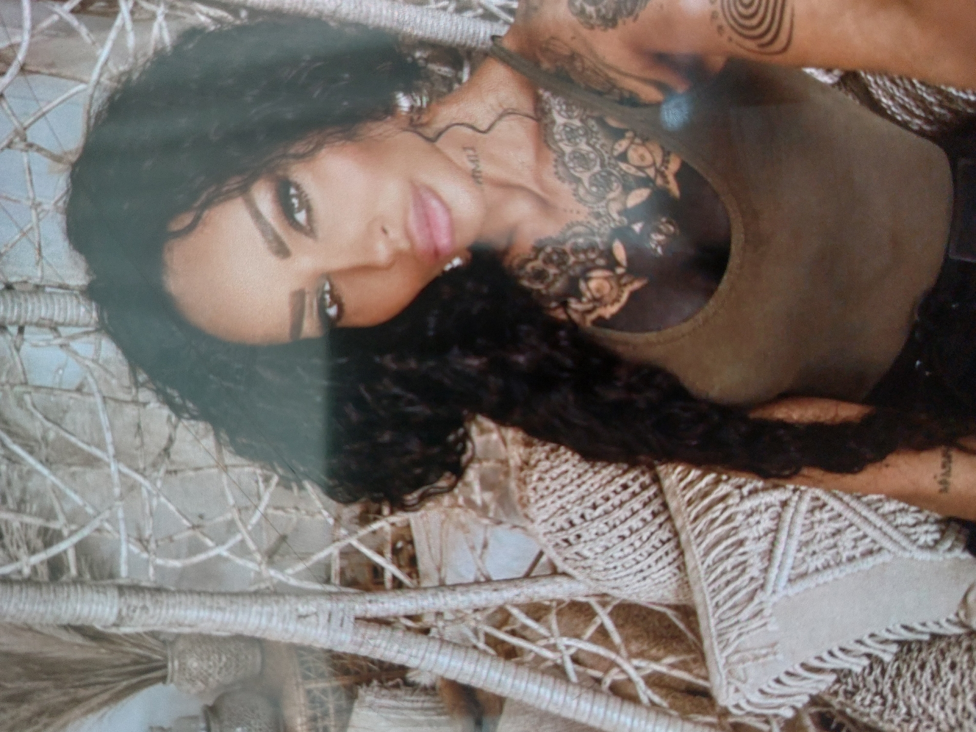 A medium shot depicts a woman with curly dark hair, lying in a white woven chair or hammock-like structure. She is wearing an olive green tank top, exposing tattoos on her chest, arms, and neck. She has light skin, dark eyes, and full lips. The woven chair has intricate patterns and textures. The background is indistinct and appears to be outdoors. The location and city of Tsarevo, Bulgaria, cannot be determined from the image.