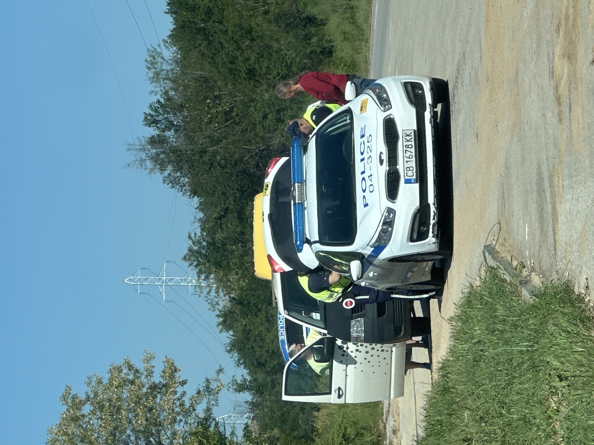 A police checkpoint scene on the outskirts of Ledenik, Bulgaria. Two police vehicles are parked on a rural roadside. Two police officers are interacting with a civilian near a white police car. A second, more heavily marked police van is parked behind. The license plate of the white car is CB 1678 KK. Power lines are visible above the trees in the background.