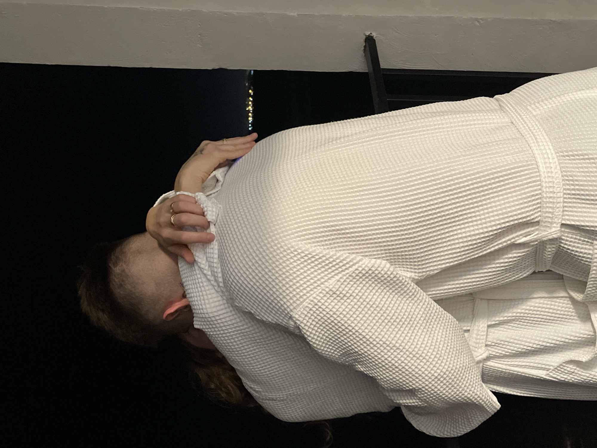 A person wearing a white waffle-textured robe is seen in an interior setting, possibly a bathroom. The person is bent forward, with one hand near their head. A hand with rings is visible. The background is mostly dark, with a ceiling and what appears to be a ladder visible above. There is a string of lights visible near the top of the image. The location is Unknown, Unknown.