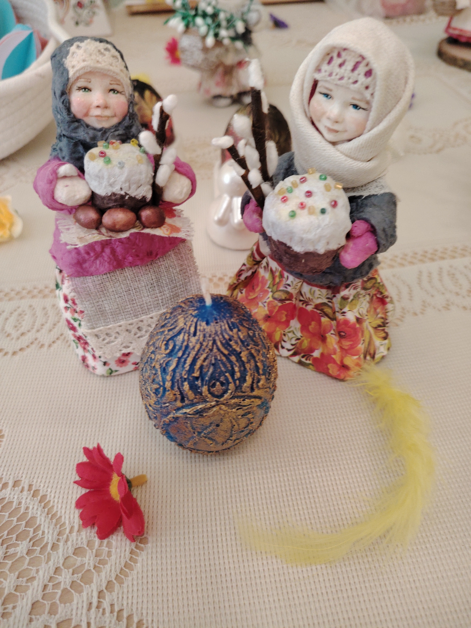 Two handcrafted dolls stand on a textured off-white tablecloth. Each doll is dressed in traditional Russian clothing, holding a miniature Easter cake decorated with colorful sprinkles. A blue and gold decorated egg-shaped candle sits in front of the dolls, flanked by a red flower and a yellow feather. The location is in Sarapul, Russia.