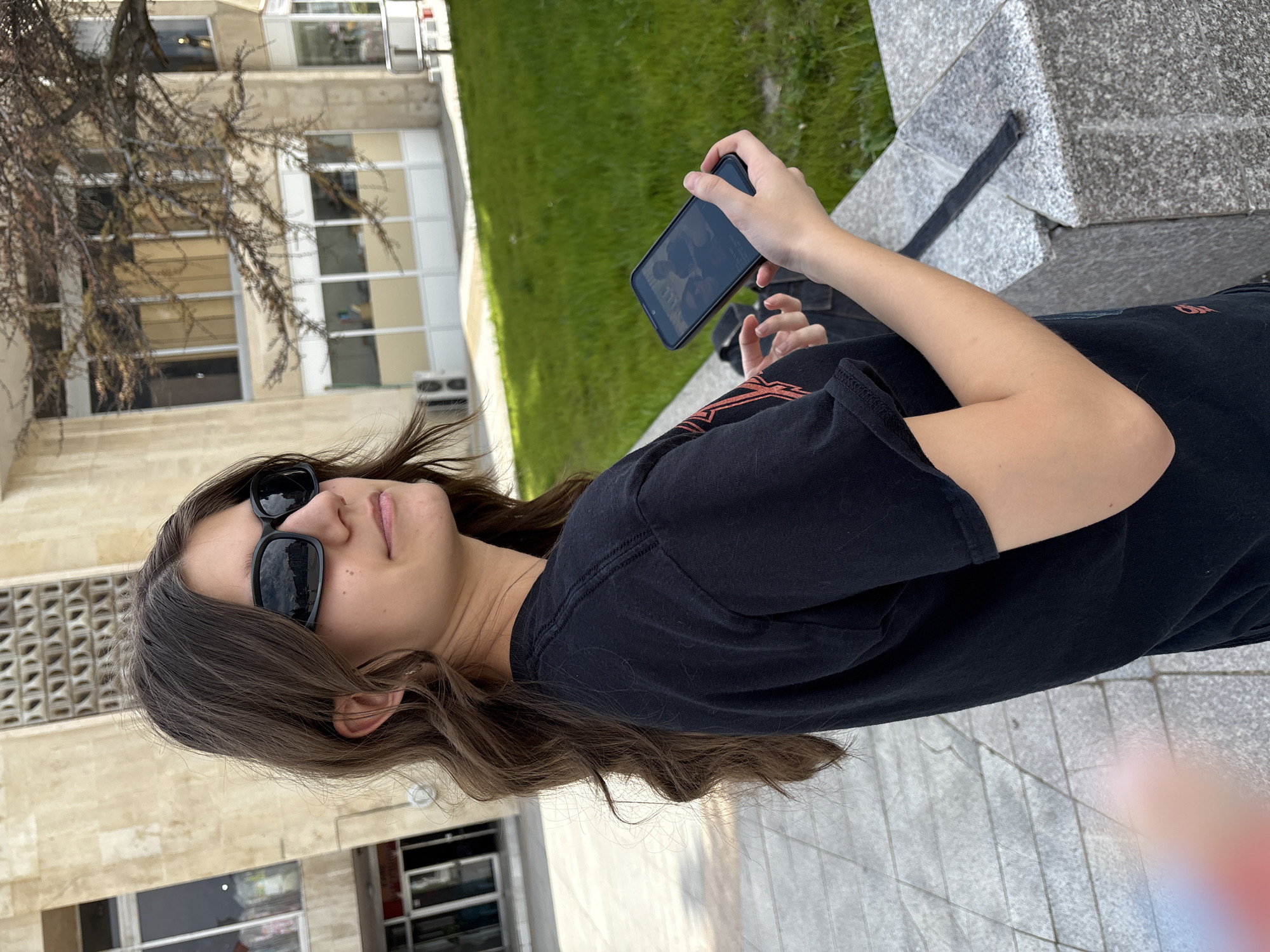 A young person with shoulder-length brown hair and sunglasses is holding a smartphone in their left hand, looking at the screen. They are wearing a black t-shirt. The setting appears to be outdoors, adjacent to a stone structure and a patch of green grass. In the background, buildings with windows and architectural details are visible. The location is potentially in Panagyurishte, Bulgaria, based on the query context.