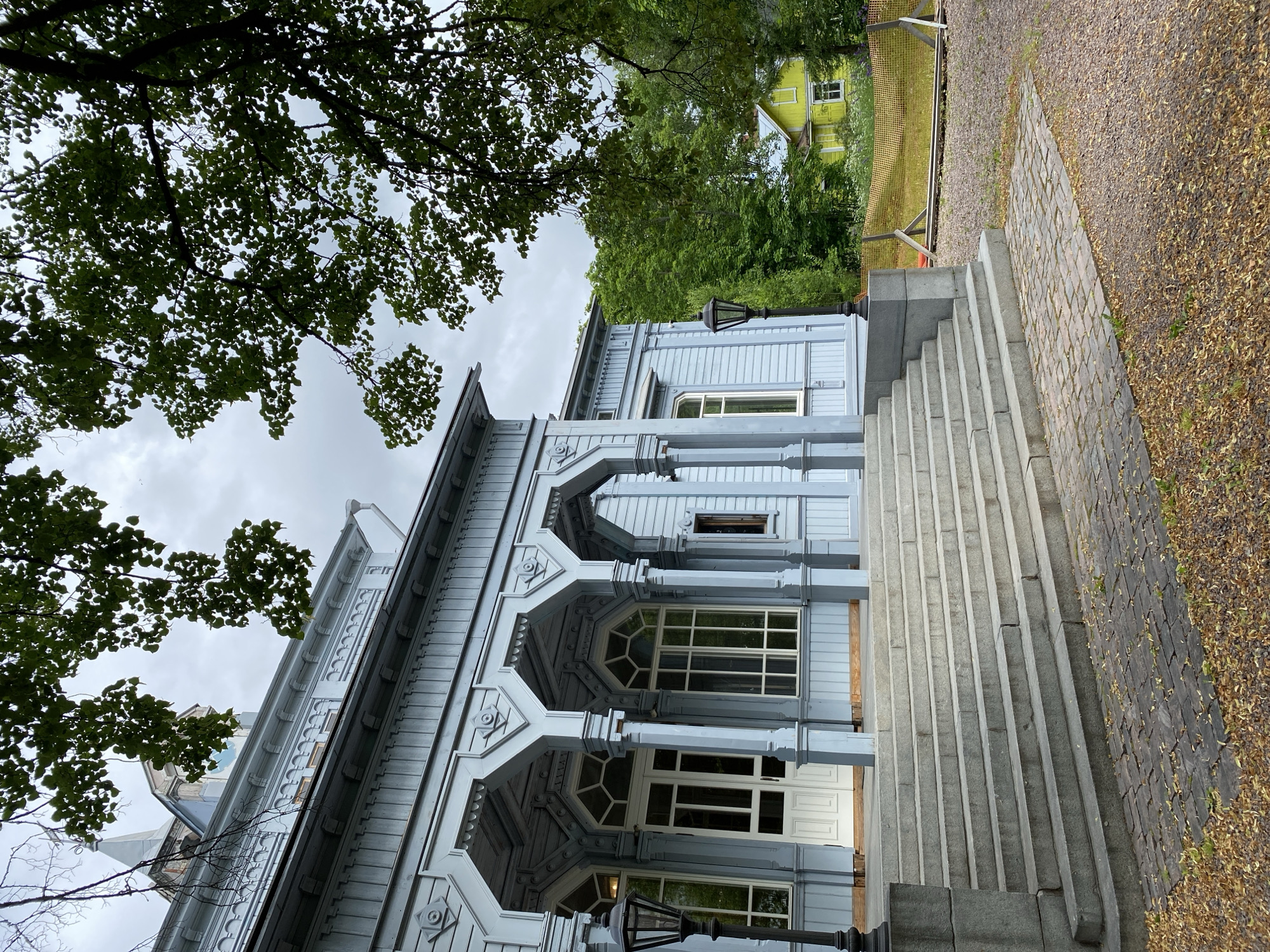 The image shows a pale blue wooden building with a covered porch supported by columns. The porch has arched openings and decorative trim. A wide set of stone stairs leads up to the entrance. To the right of the building, there is a bright yellow house visible through the trees, and a section of orange fencing. The ground in front of the stairs is paved with cobblestones and covered with fallen leaves. Trees with green foliage are prominent in the foreground and background. A cloudy sky is visible above. This building is the "Kantele" House, a museum in Sortavala, Russia, dedicated to the Karelian instrument and culture.