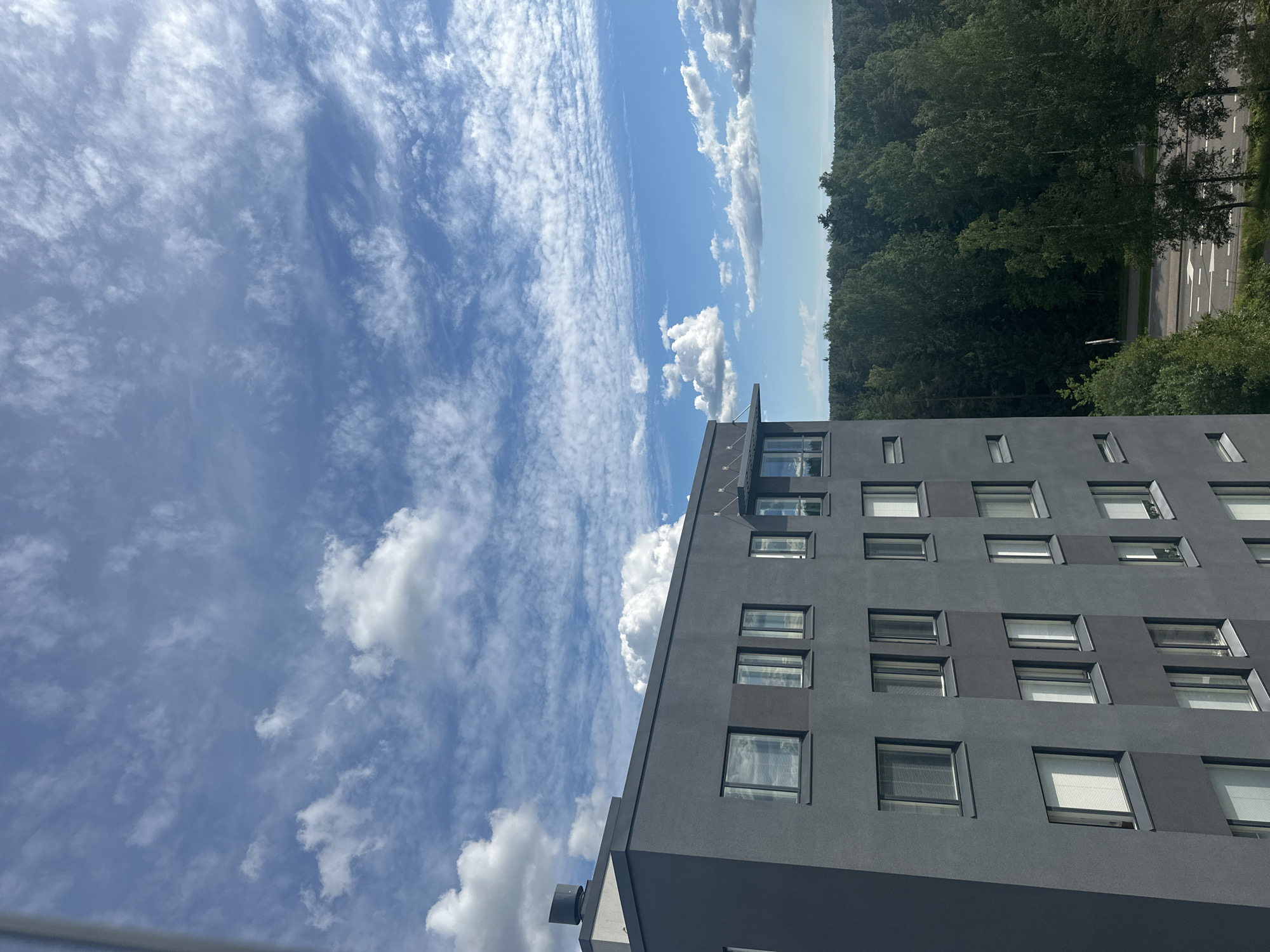 The image shows a modern, dark gray building with multiple windows, possibly an apartment complex or office building. The building is set against a backdrop of a lush, green forest. The sky is blue with scattered white clouds, some appearing thin and wispy, others more cumulus-like. A road with white lane markings is visible in the lower right portion of the image, bordered by trees. The scene appears to be a daytime shot, likely in a suburban or semi-rural area.