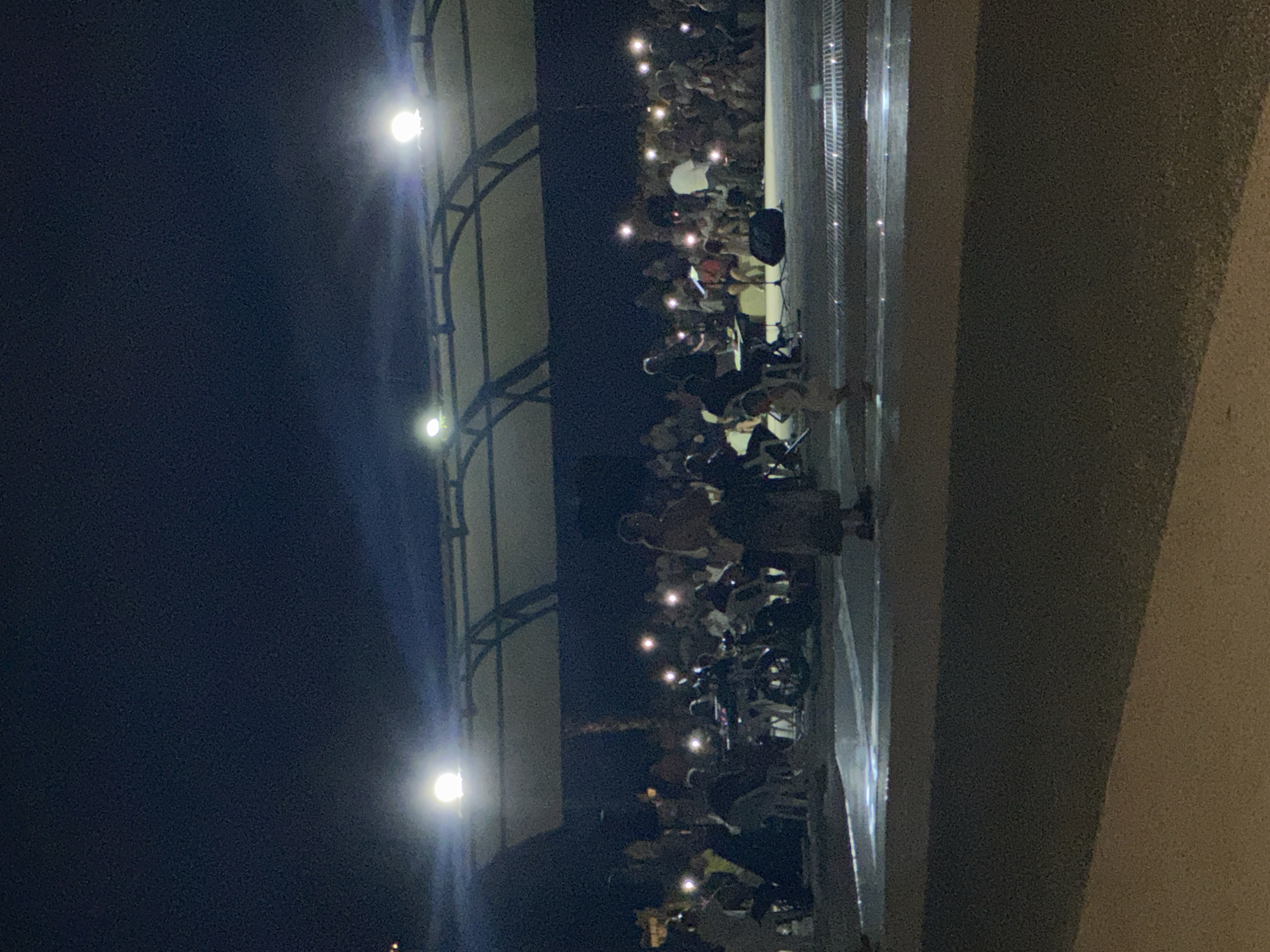 The image depicts an outdoor night scene, possibly a concert or performance, in Fethiye, Turkey. A large crowd is gathered, many illuminated by the flashlights of their mobile phones. In the foreground, a woman stands with her back to the camera, wearing a long, dark skirt and a lighter-colored top. Further back, a small group is gathered around what appears to be a musical setup, with instruments and a keyboard visible. A child in a light-colored outfit is standing in the middle of the scene, facing away from the camera. Above, a covered structure with lights suggests a stage or seating area. The overall ambiance is dimly lit, with artificial lights creating bright spots throughout the crowd and on the ground.