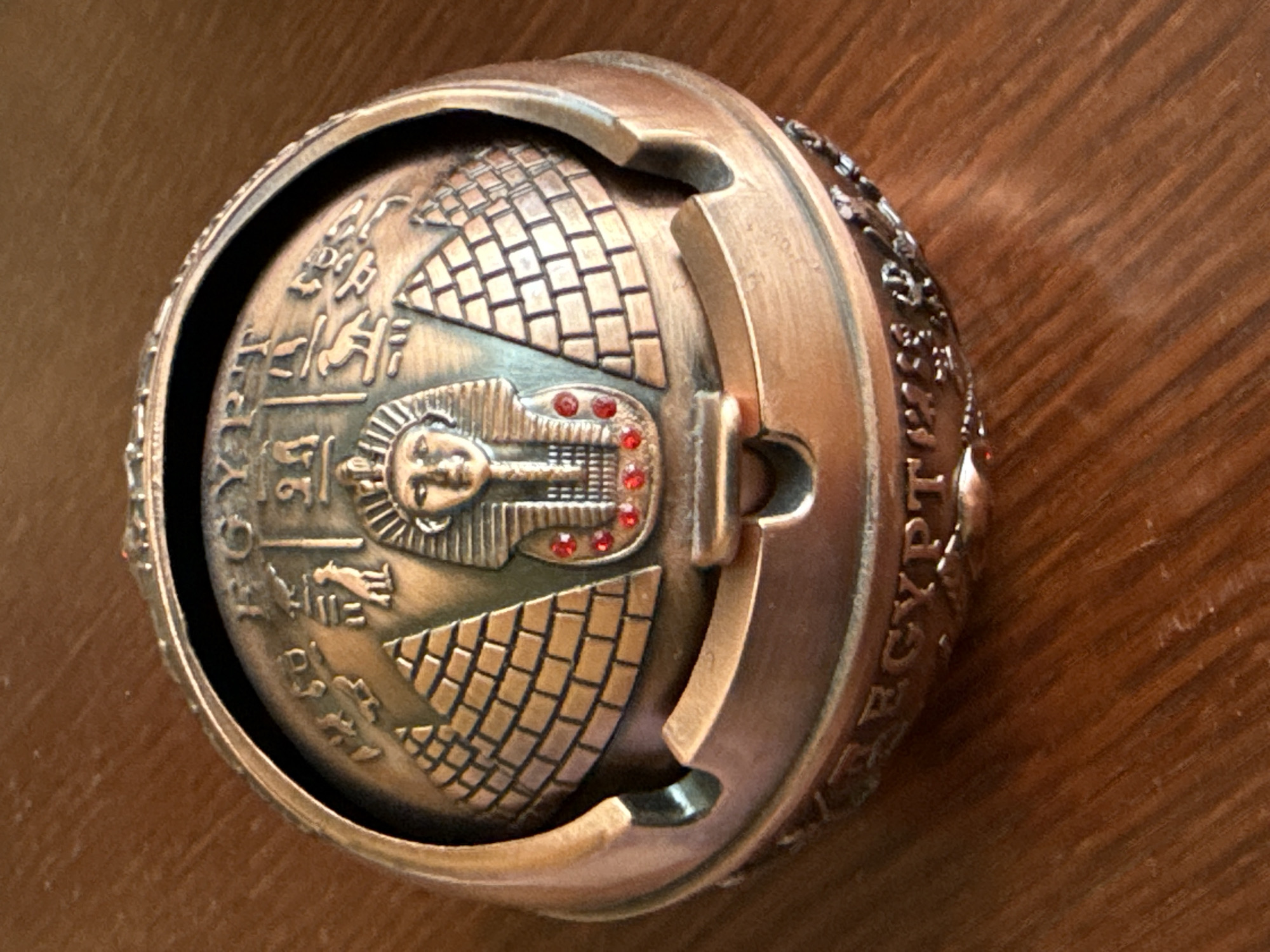This is a close-up, high-angle, indoor shot of a decorative, round, metallic object, possibly a ring or a locket, resting on a wooden surface. The object has an Egyptian theme, featuring a central carved image of a pharaoh's face wearing a nemes headdress. The pharaoh's face is detailed, with eyes, nose, and mouth visible, and is adorned with small red stones, possibly rhinestones, in place of jewels on the headdress. Flanking the pharaoh's head are stylized depictions of pyramids, rendered with brick-like patterns. Hieroglyphic symbols are carved into the metal around the central imagery.
The object appears to be made of a bronze-toned metal, with some areas showing darker patina, suggesting age or deliberate design. The outer edge of the object is also decorated with relief carvings, and the word "EGYPT" is partially visible in raised lettering on the right side, confirming the theme. The wooden surface it rests on has a warm, brown tone with a visible wood grain pattern, indicating it is likely a table or desk. The lighting is warm and seems to come from above and the left, casting subtle shadows on the object and highlighting its textured surfaces. No specific activity or event is taking place; the object is simply displayed. The overall impression is one of an artifact or collectible with an ancient Egyptian motif.
