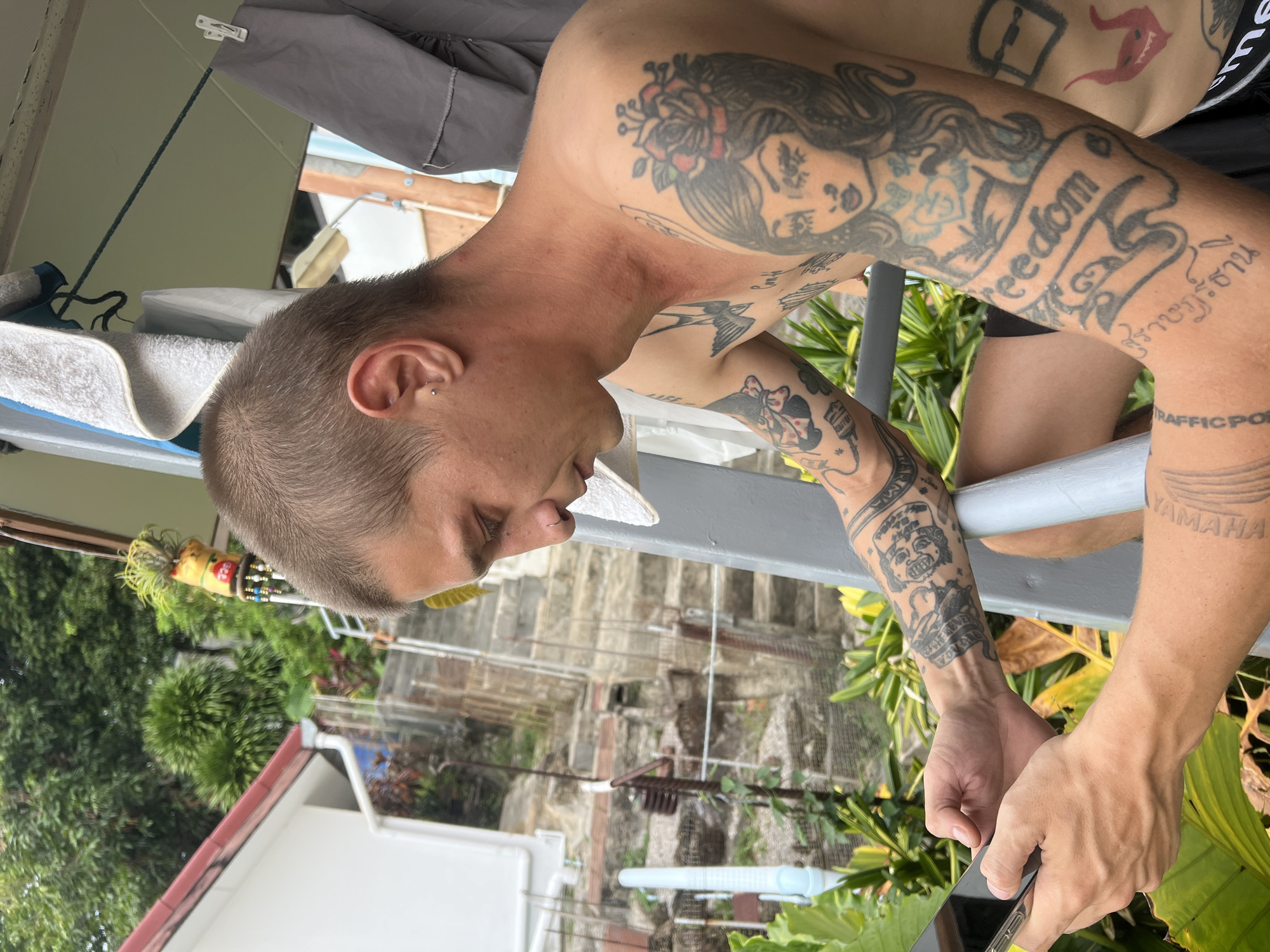 A shirtless man with short brown hair, an earring, and multiple tattoos on his arms and chest sits outside and looks down at his phone. He is wearing dark shorts. In the background, there is a white building with a red roof, a concrete staircase, lush green foliage, and some laundry hanging on a line. The man is holding a smartphone in his left hand and appears to be typing or scrolling.