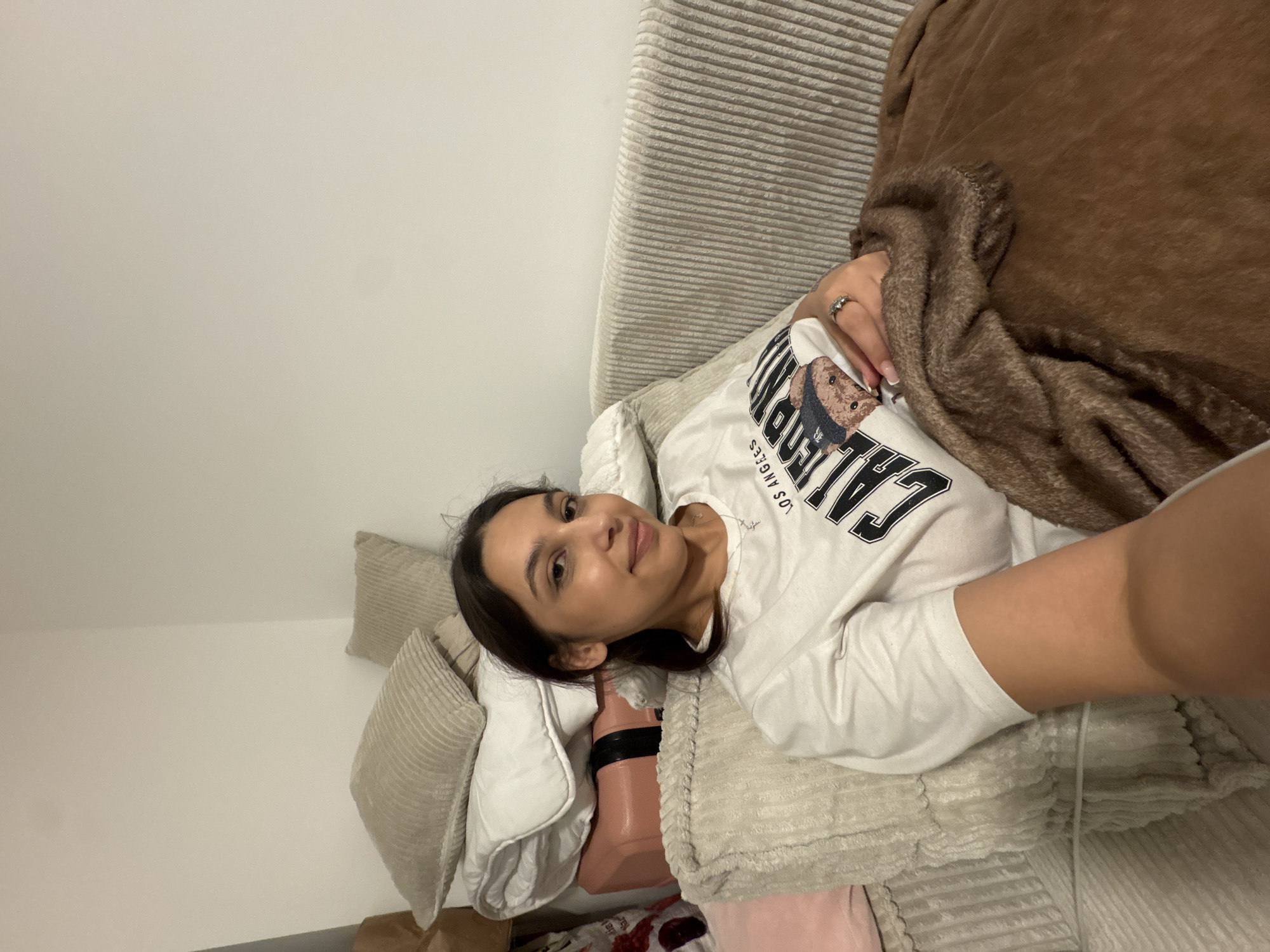 A young adult female is seated on a light beige corduroy sofa in Mönchengladbach, Germany, gazing directly at the viewer with a subtle smile. Her dark hair is pulled back, and she wears a white short-sleeved t-shirt featuring "LOS ANGELES CALIFORNIA" text and a graphic of a bear. A silver-toned ring is visible on the ring finger of her right hand, which rests on a soft, brown plush blanket covering her lower body. Behind her, stacked against a plain, light-colored wall, are several light-colored corduroy pillows, folded white bedding, and a pink hard-sided case with a black strap. Additional obscured items are visible to the far left. The environment appears to be an indoor residential setting with ambient lighting.