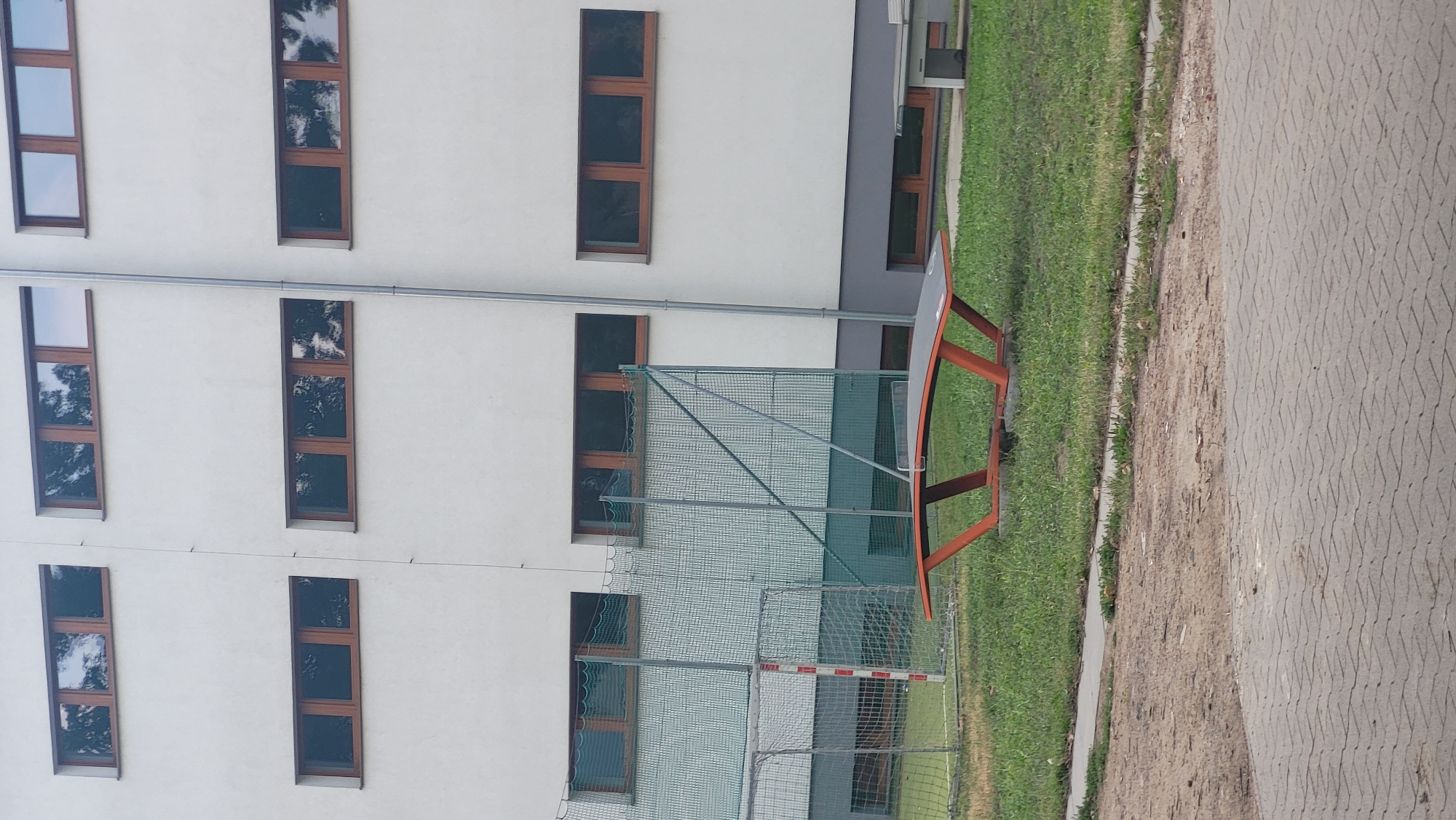 The image depicts an outdoor scene in the city of Prešov, Slovakia. In the foreground, there is a paved walkway made of interlocking concrete pavers. Behind this, a grassy area is visible, and on the grass, a bright orange and gray ping pong table is centrally placed. Further back, a chain-link fence with a green mesh overlay encloses a sports field, with a white goalpost partially visible. The background is dominated by a modern, white building with multiple horizontal windows featuring dark frames. The windows reflect the surrounding trees and sky. The building appears to be multi-story, with at least three visible rows of windows. The overall impression is of an educational or recreational facility.