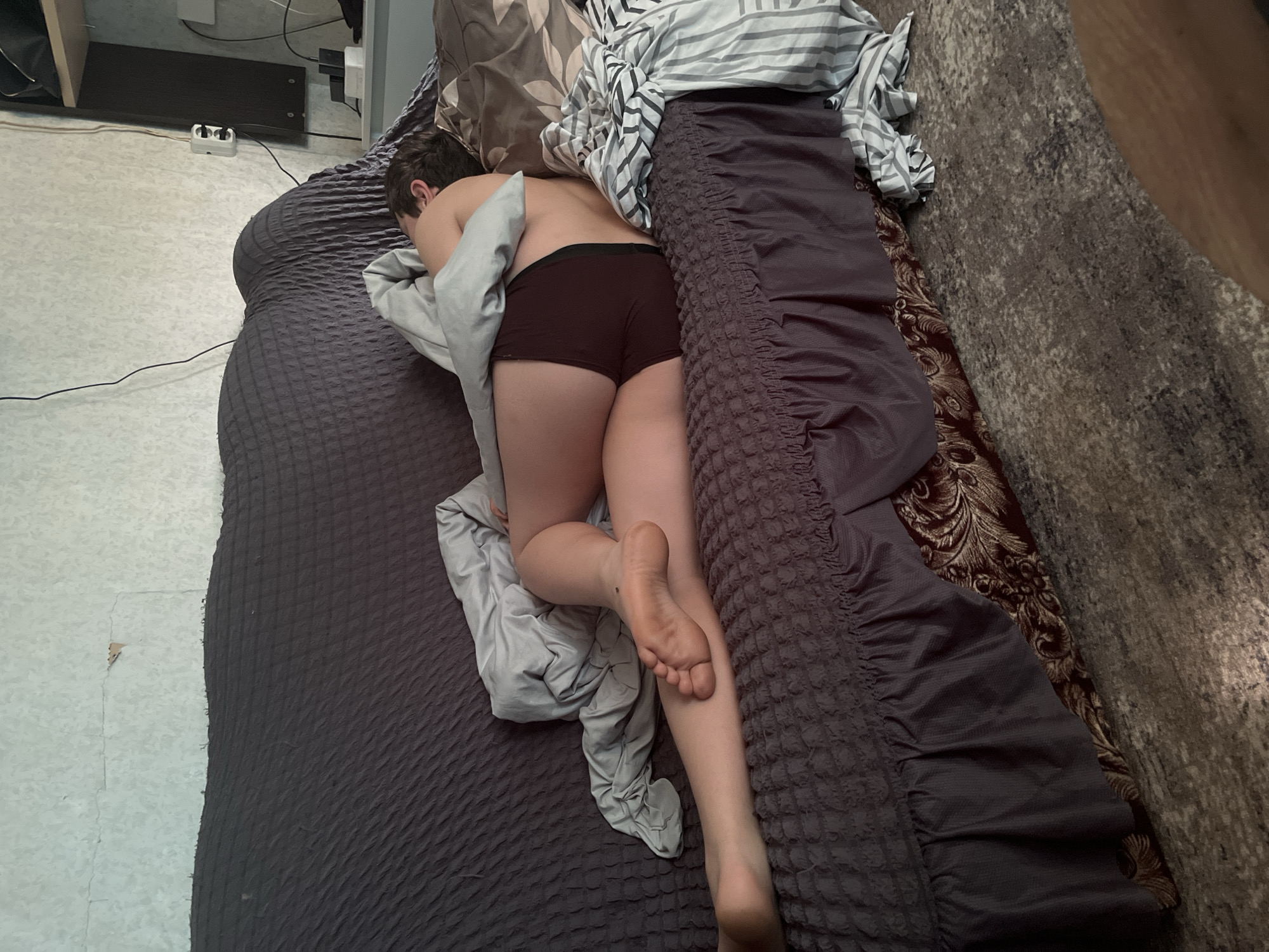 An overhead shot depicts an individual resting face down on a bed in an indoor setting, likely a bedroom in Фоки, Russia. The person, who appears to be an adult or young adult, is wearing dark-colored boxer briefs or shorts. Their head is turned away from the camera, and their dark brown hair is visible. A light grey blanket or sheet is partially covering their upper body, while their lower back, buttocks, and legs are exposed. The sole of one foot, showing a small mole, faces upwards, appearing slightly soiled.
The bed is covered with a dark grey, textured comforter. Beneath a striped pillow or sheet near the person's head, a brown pillow with an ornate gold pattern is visible. To the left of the bed, a light-colored tiled or linoleum floor can be seen, where a white power strip or adapter with several cables is plugged into an outlet near a wall. Dark cables extend across the floor. To the right of the bed, a dark, textured rug covers the floor. The lighting is ambient, without strong shadows, making the precise time of day difficult to determine. No text is visible in the image. The scene suggests a moment of rest or sleep in a domestic environment.