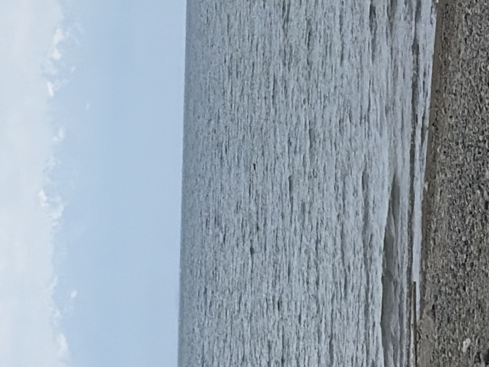 A serene, slightly overcast day unfolds at the edge of a vast body of water, likely a lake or ocean. In the foreground, a pebble beach stretches across the bottom of the frame, its grey and brown stones varying in size. Gentle waves lap at the shore, creating a thin, wet line where the water meets the land. The water itself is a cool, steely blue-grey, with a rhythmic pattern of ripples and small waves extending towards the horizon. In the far distance, a mountain range rises, its peaks capped with white snow, forming a hazy, majestic backdrop against the pale blue sky. The overall atmosphere is one of quiet natural beauty.