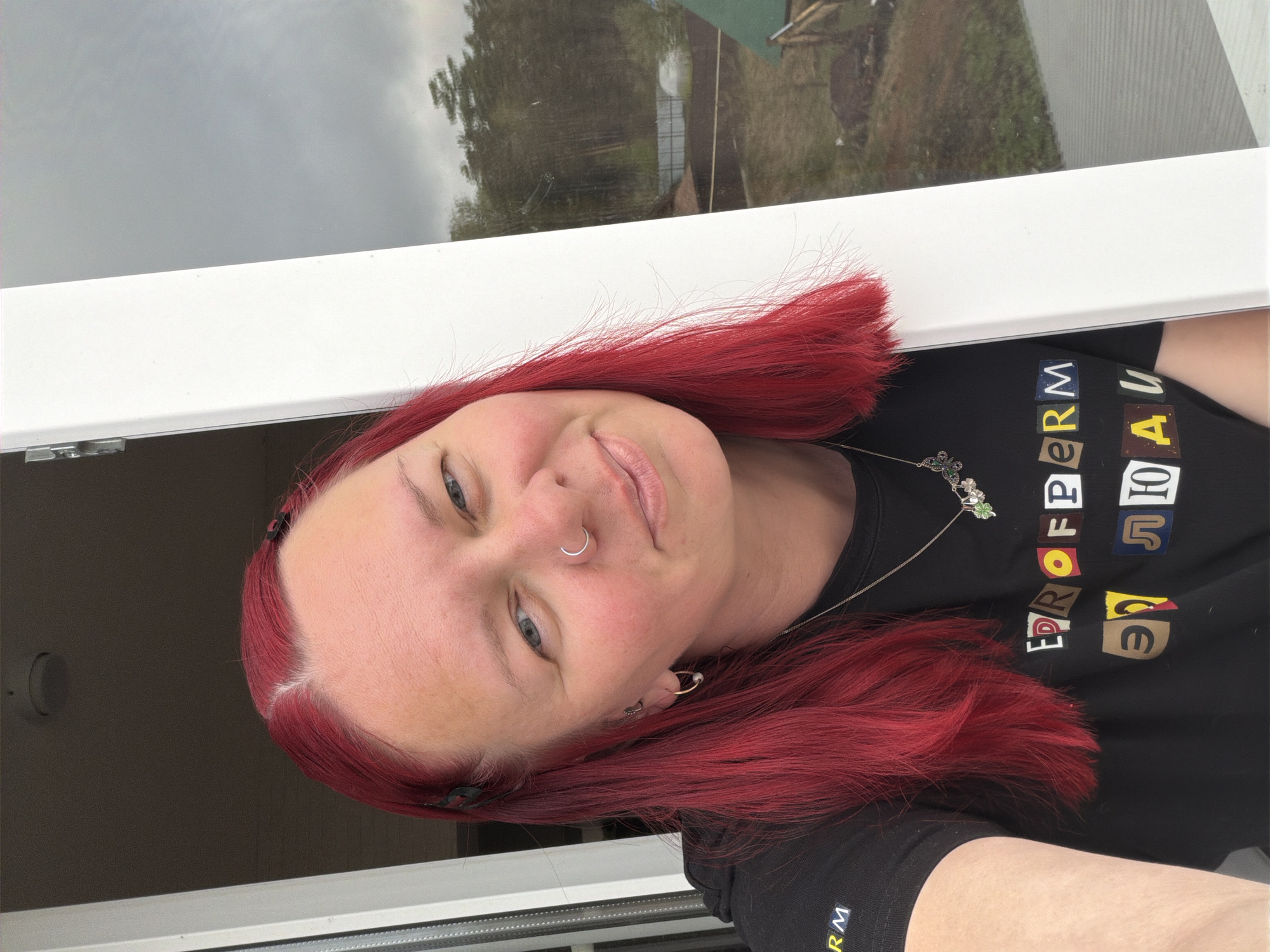 A woman with bright red hair stands by an open window. She has fair skin and is wearing a nose ring. She is wearing a black t-shirt with colorful lettering. Through the window, trees and buildings are visible. The city of Chaikovsky, Russia is not explicitly verifiable from the image provided.