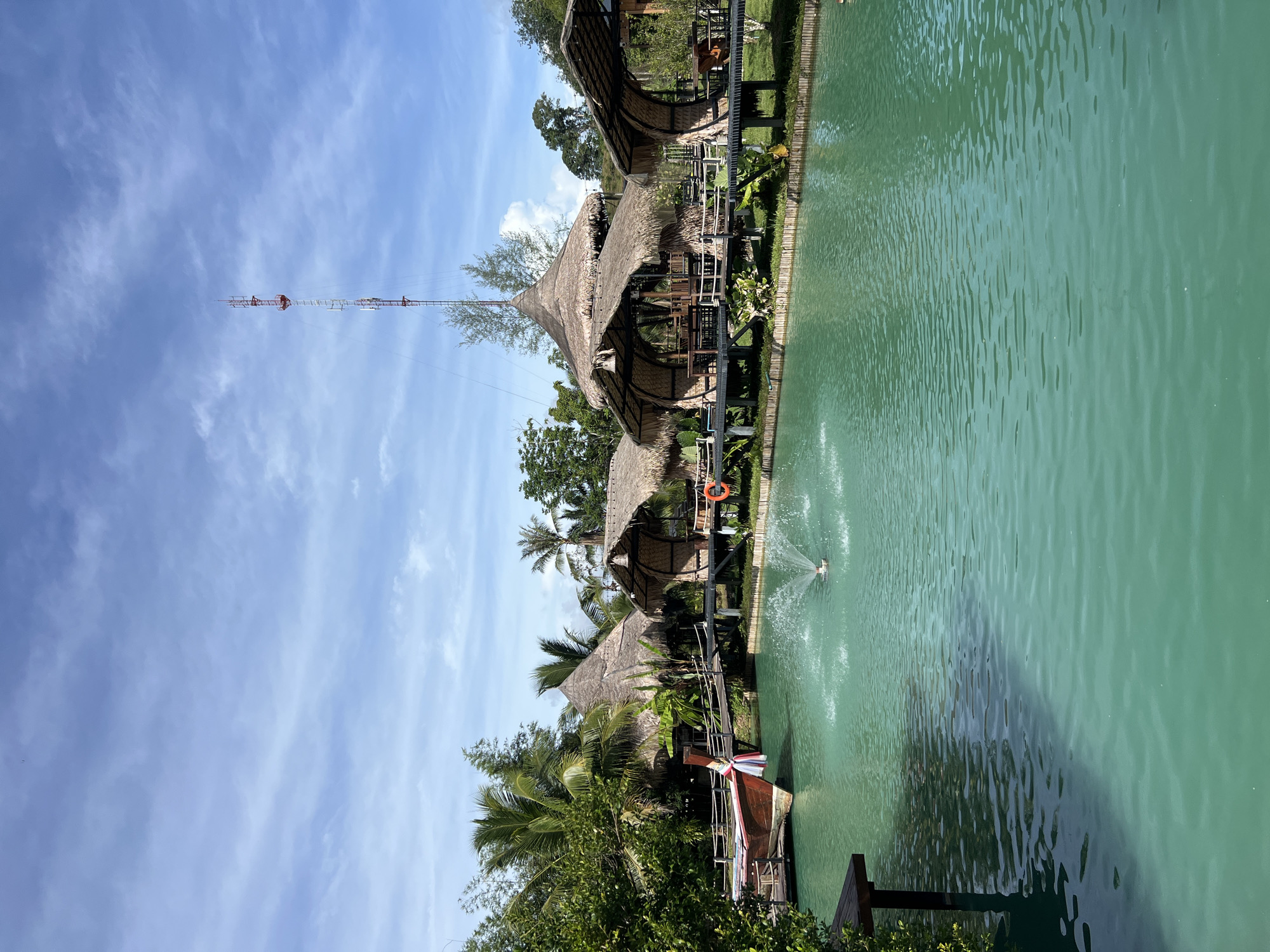 The image depicts a tranquil waterfront scene in Tambon Pa Kae Bo Hin, Thailand. A vibrant turquoise body of water dominates the foreground, disturbed by a central fountain spraying water upwards. To the left, a traditional Thai longtail boat with a red hull and wooden accents is docked, adorned with a colorful ribbon.
A wooden walkway spans the water, connecting to several thatched-roof huts or pavilions built on stilts. These structures, with their natural, rustic aesthetic, appear to be part of a resort or restaurant. Palm trees and lush green vegetation surround the huts, framing the scene with tropical foliage. In the background, a tall communication tower rises against a bright blue sky dotted with wispy white clouds. The overall atmosphere is one of serene, tropical beauty.