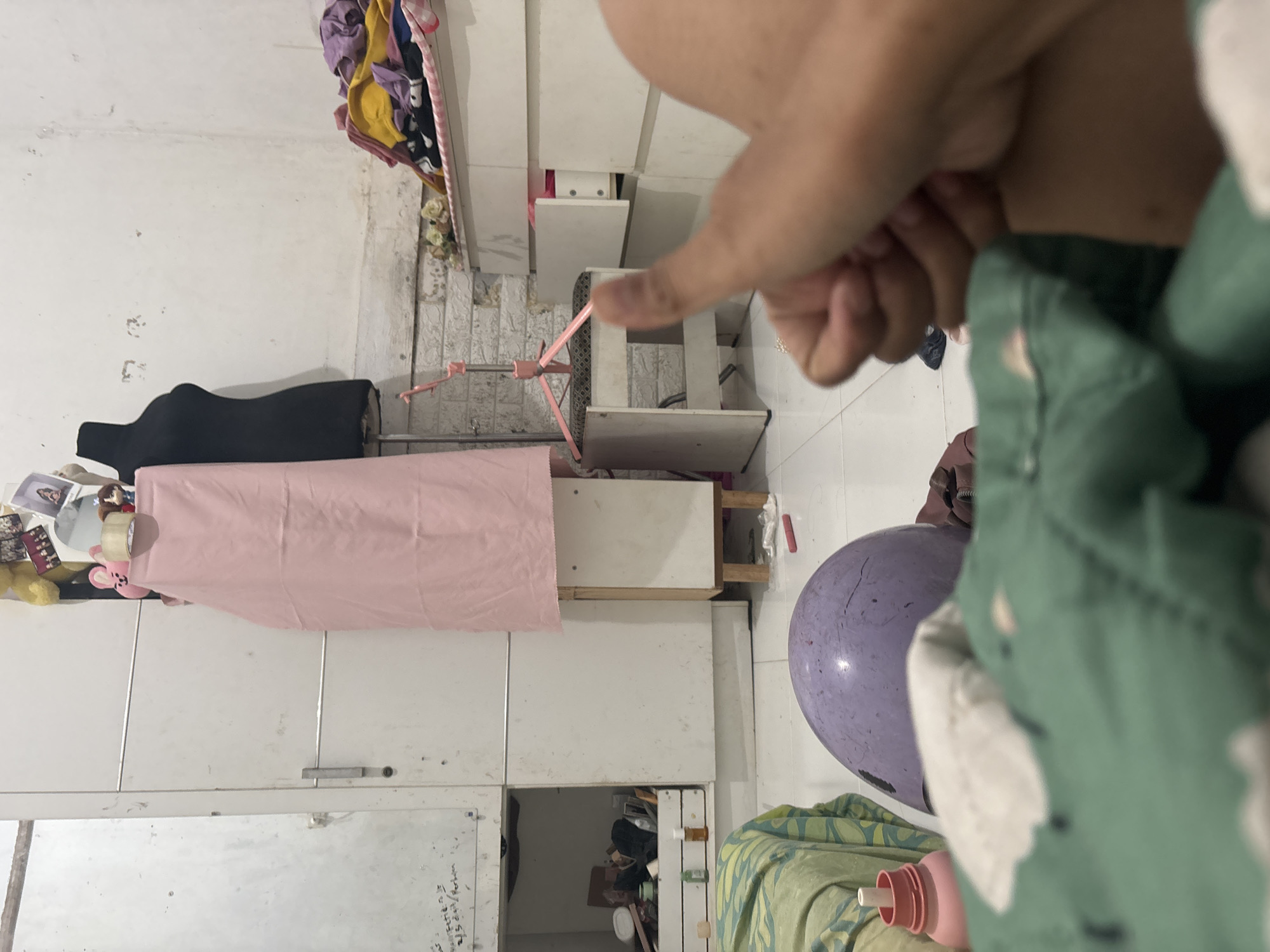 This image depicts an indoor scene, likely a bedroom or storage area, with a slightly cluttered and lived-in appearance. A person's hand giving a thumbs-up gesture is prominently featured in the foreground, out of focus. In the middle ground, a purple helmet rests on a green, patterned fabric, possibly a blanket or clothing. Behind these items, a white wardrobe is visible on the left, with an open compartment revealing various objects. To the right, a black mannequin stands beside a pink fabric draped over a stand. Further to the right, white drawers are stacked, with one drawer partially open showing colorful items. The background shows a white wall with some discoloration and what appears to be exposed brickwork near the floor. The overall lighting is dim. There is no explicit indication of the location being North Jakarta, Indonesia; this is an external context provided.