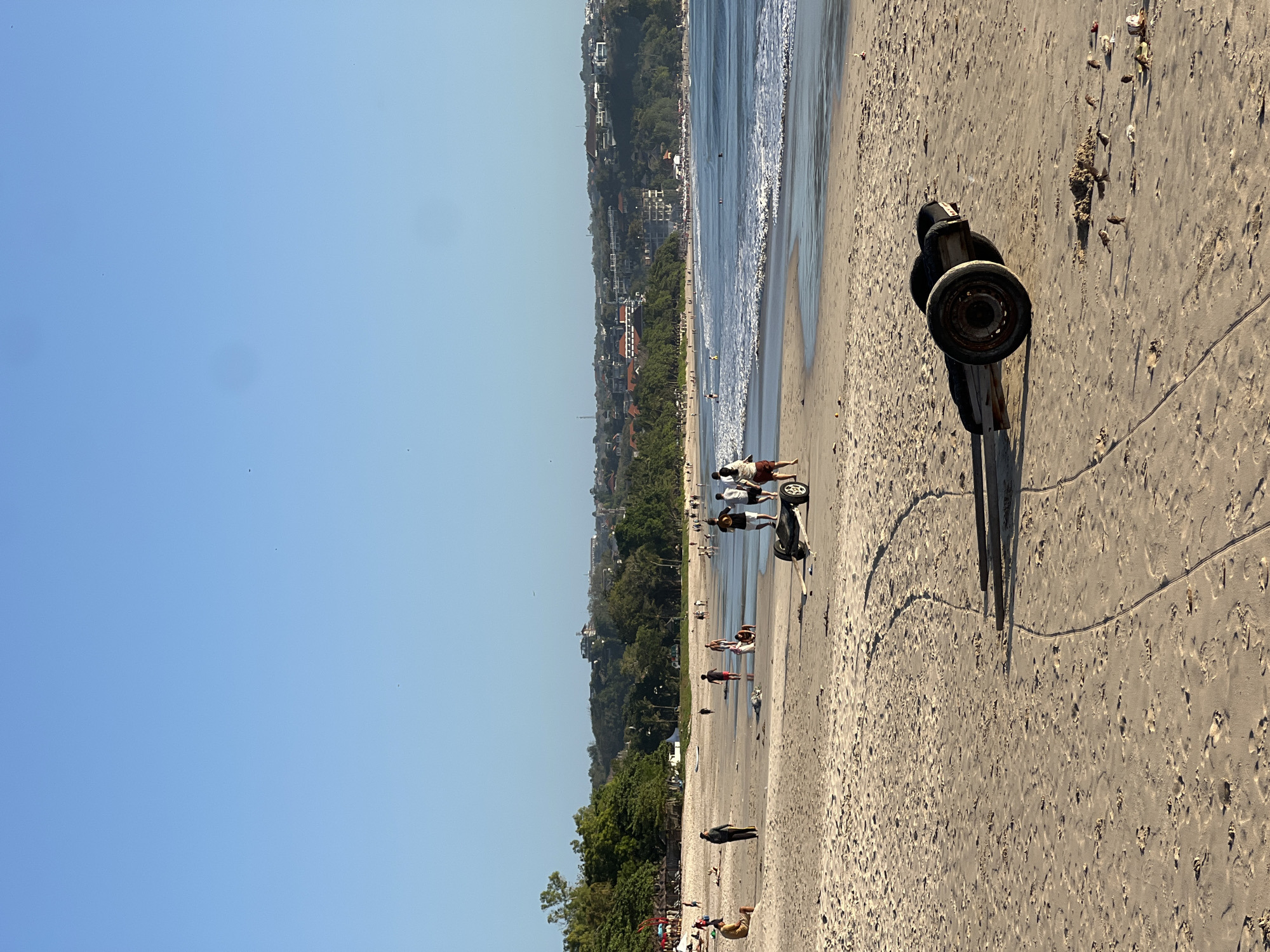 The image depicts a beach scene in Kuta Selatan, Indonesia. In the foreground, a weathered trailer with two wheels sits on the sand. Tire tracks are visible on the sand leading away from the trailer. A flock of small birds is pecking at something on the sand near the trailer.
Further into the scene, people are walking along the shoreline and in the shallow water. Some are casually strolling, while others appear to be engaged in activities such as playing in the water or collecting items. The ocean is calm with gentle waves breaking on the beach.
In the background, lush green trees and vegetation line the coast, with buildings and structures visible on the hillside overlooking the beach. The sky is clear and blue, indicating good weather. The overall scene conveys a relaxed atmosphere of a popular beach destination.