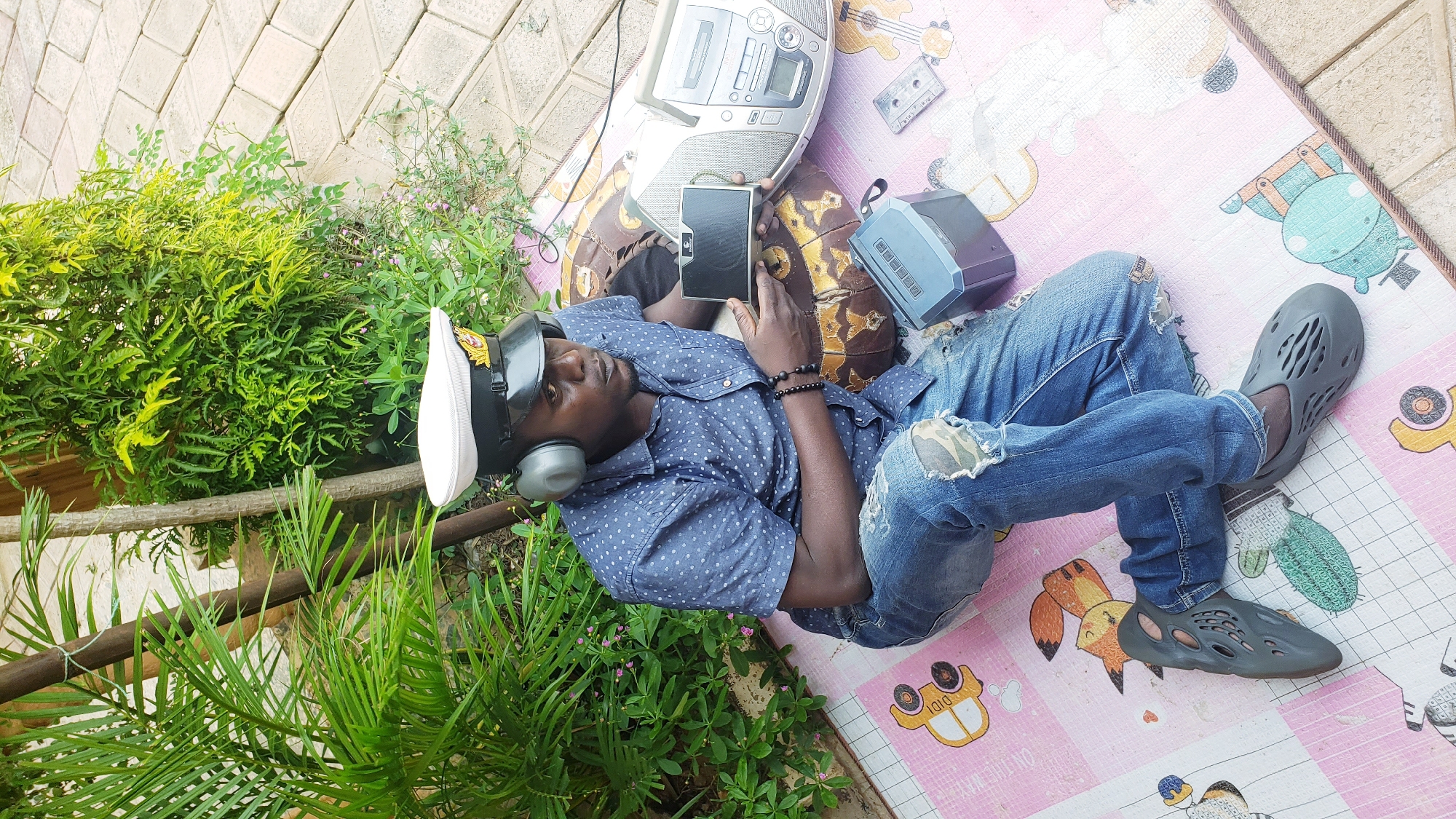 In Narayi, Nigeria, a Black man wearing a blue polka-dot shirt, distressed denim jeans, grey clogs, and a white captain's hat with headphones sits on a mat outdoors. He holds a black tablet and appears to be interacting with it. Beside him are a silver boombox, a grey speaker, and a cassette tape. The background features lush green foliage and a paved walkway.
