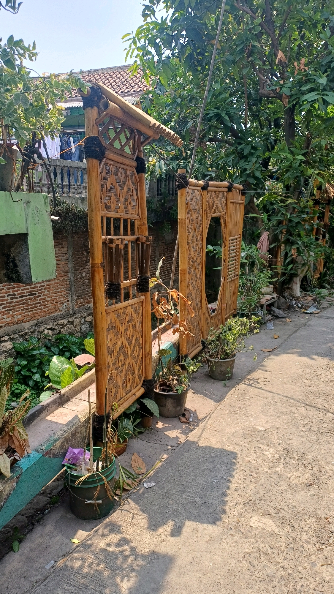In Citeureup, Indonesia, an outdoor scene unfolds under a bright, clear sky. The foreground is dominated by a pathway of rough, grey concrete, casting long shadows from the surrounding objects. To the left, a low wall, painted a faded turquoise green, separates the path from a raised garden bed. This bed is filled with lush green foliage and some dried leaves.
The prominent feature of the scene is a decorative entryway constructed entirely from bamboo. The entryway consists of two upright panels woven from bamboo slats, creating intricate patterns. The left panel is wider and features a small, rectangular window with vertical bamboo slats. It is topped with a decorative bamboo roof structure. The right panel is narrower and has a large, irregular oval opening. Both panels are supported by thick bamboo posts. Black rope is used to bind the bamboo elements together.
Several potted plants are placed around the bamboo entryway. Two dark grey pots, one in front of the right panel and another to the far right, contain various green plants. A dark green bucket, positioned near the left panel, holds dried plant stems and a crumpled purple plastic bag.
Behind the bamboo entryway, a brick wall, partially covered by green paint on the left side, forms a backdrop. Further back, a tiled roof of a building is visible, along with clothes hanging on a clothesline. Trees with dense green foliage tower over the scene, creating a natural canopy. The overall impression is of a rustic, outdoor space with elements of traditional Indonesian craftsmanship.