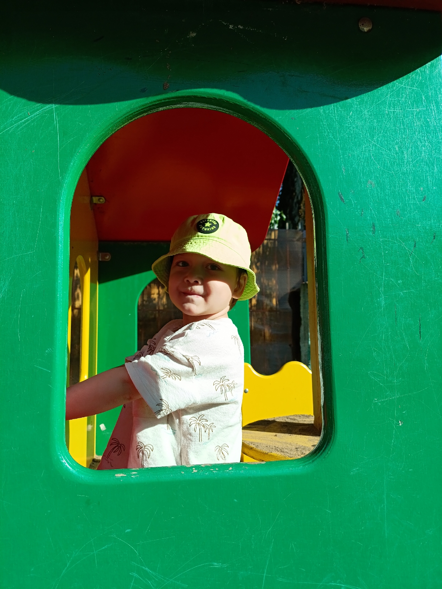 A young child wearing a pale yellow bucket hat and a light-colored t-shirt with palm tree patterns is framed within a green, red, and yellow structure, likely a playhouse or part of playground equipment. The child is looking at the camera with a slight smile. The structure appears to be in an outdoor environment with visible foliage and shadows in the background. The green surface of the structure has visible scratches and wear. Based on the provided information, the scene is located in the city of Chaikovsky, Russia.