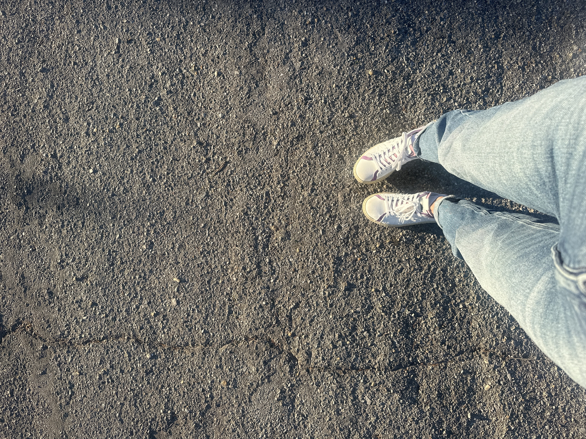 An overhead shot shows a person's legs and feet standing on a rough, asphalt surface. The person is wearing light-wash denim jeans and white sneakers with purple accents and laces. The asphalt is textured with small stones and shows a crack running vertically down the left side of the frame. The lighting suggests it is daytime with sunlight casting shadows on the ground. The image does not provide enough information to determine a specific location in the city of Shumen, Bulgaria.