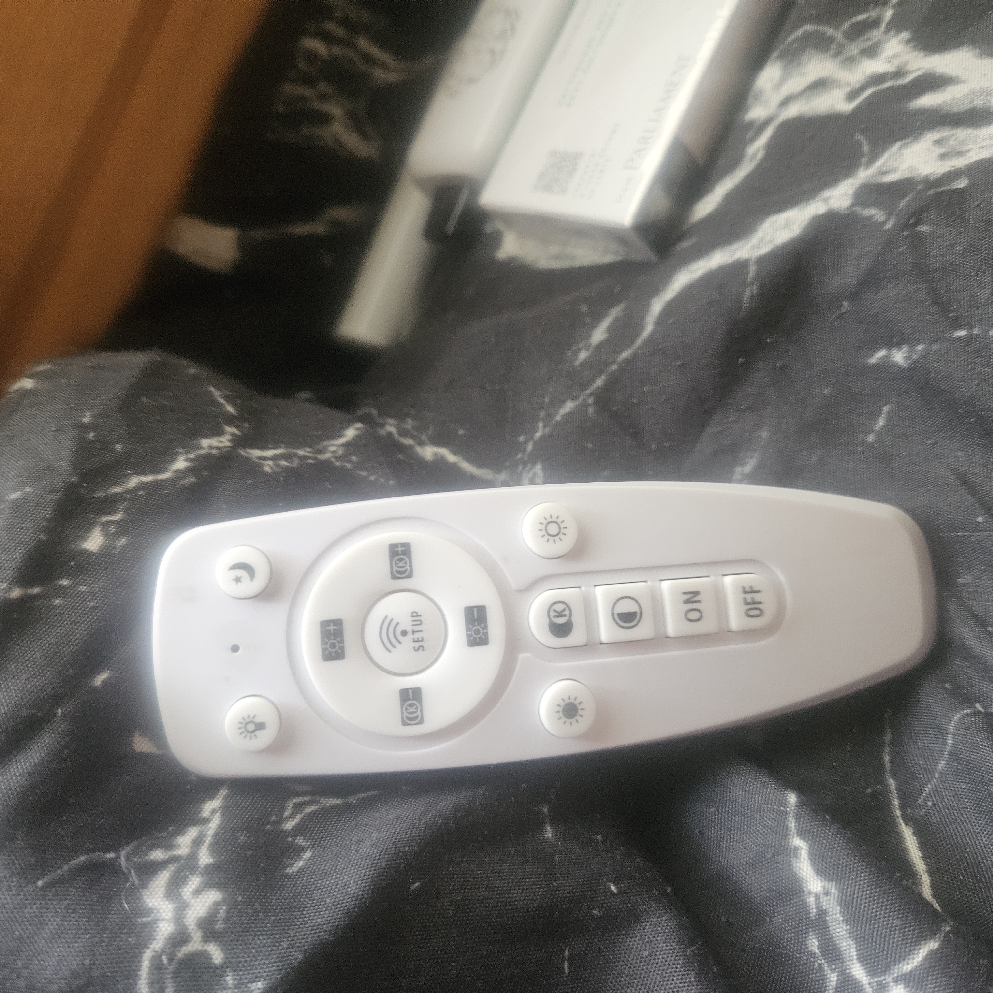 This is a medium-angle, close-up shot of a white remote control with various buttons, resting on a black fabric with a marbled pattern. The remote is the primary object of focus.
The remote control has several buttons, some circular and some rectangular. The circular buttons feature icons: a crescent moon with a star, a sun with rays, and a lightbulb. There are also plus and minus symbols within circles, suggesting brightness or intensity adjustments. A larger, central circular button is labeled "SETUP" and has a wavy antenna icon above it. Rectangular buttons are labeled "ON" and "OFF." Other circular buttons feature a "K" icon and a lightbulb icon with rays.
The fabric it rests on is dark, likely black, with white or light gray streaks that resemble a marble pattern. The lighting in the scene appears to be indoor, and the time of day and weather are not discernible. There are no people present in the image.
In the background, slightly out of focus, are parts of white objects, one of which appears to be a rectangular box with some text on it. The visible text includes "PARLIAMENT" and a QR code.
The overall impression is that of a functional item in a domestic or personal setting. The image seems to be taken in a casual manner, with the remote as the subject of interest.