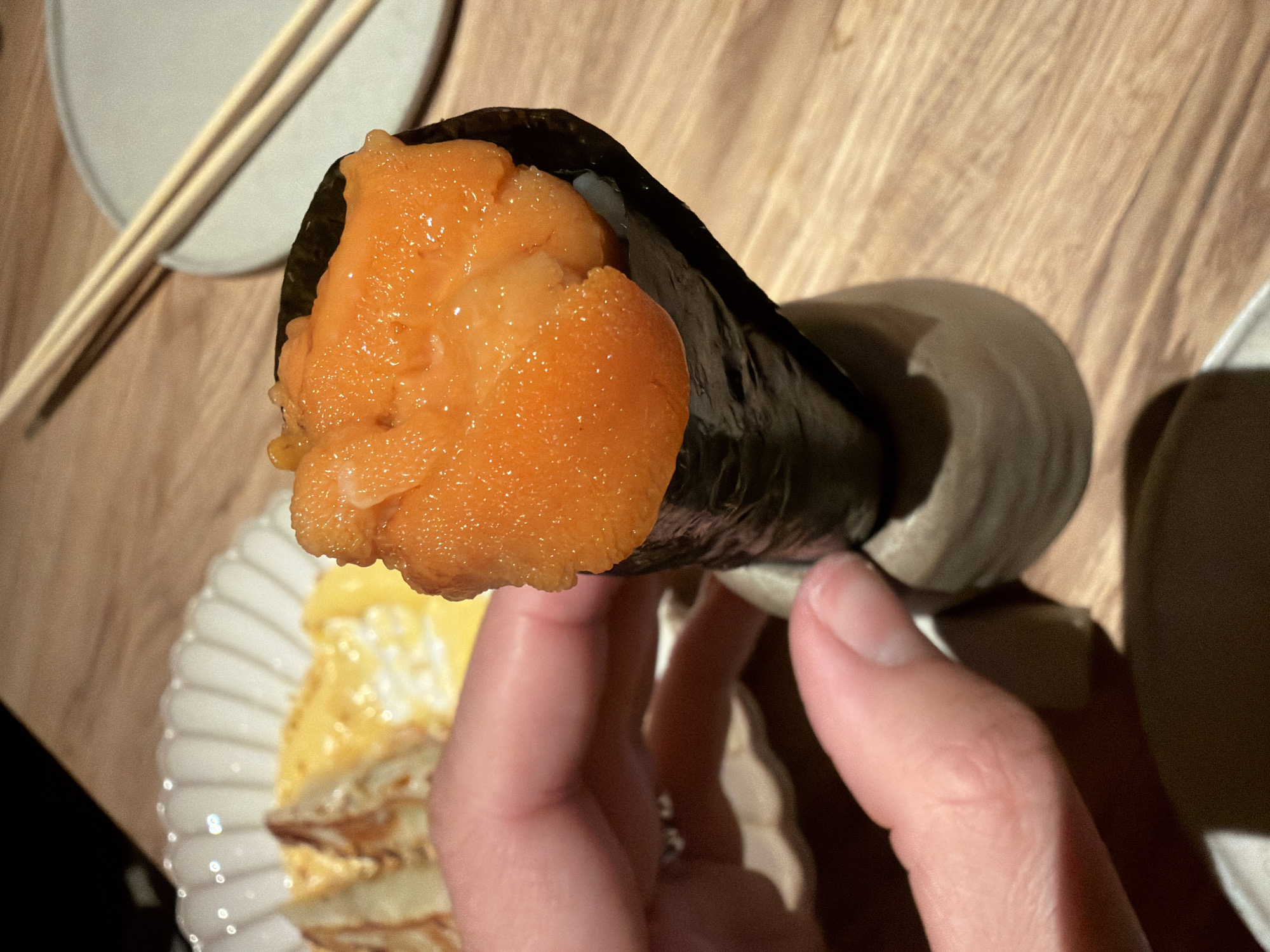 The image displays a close-up view of a person's hand holding a temaki (hand roll) filled with bright orange uni (sea urchin roe) in an indoor dining setting, likely in Moscow, Russia. The temaki, wrapped in dark nori (seaweed), is generously packed with the glistening, fresh uni.
The hand, with visible fingers and thumb, firmly grasps the temaki. In the blurred background to the left, a light-colored, ridged ceramic plate holds a yellowish, layered dish, possibly tamagoyaki or a similar culinary item. Above this plate, a pair of light-colored chopsticks rests on a small, round, greyish plate. A ceramic holder, possibly for the temaki, is partially visible to the right, blending into the shadows.
The scene takes place on a light-colored wooden table, identifiable by its grain patterns. The lighting is warm and somewhat dim, creating a cozy atmosphere, likely during an evening meal. Shadows are cast by the objects, adding depth to the composition. No text is visible in the image. The overall impression is one of a delicious and carefully presented meal.