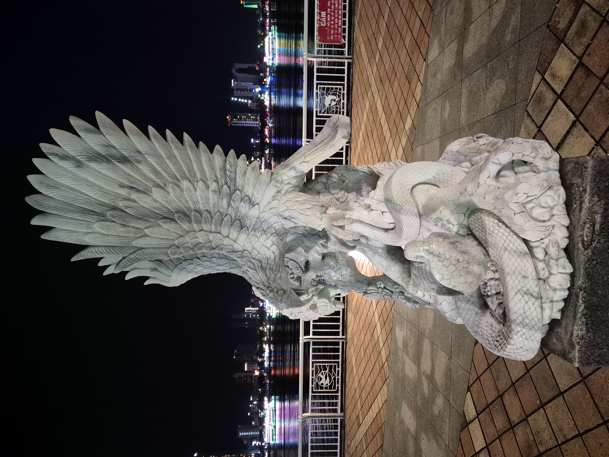 This is a nighttime, eye-level, full-shot photograph of a white stone sculpture depicting an eagle in combat with a serpent, located in the city of Đà Nẵng, Vietnam. The sculpture is positioned outdoors on a paved walkway adjacent to a body of water. The eagle, with its wings spread wide and beak open, appears to be attacking the serpent, which is coiled around the eagle's body. The sculpture is detailed, with individual feathers and scales clearly defined.
In the background, the city skyline is illuminated by numerous lights. Buildings of various heights are visible, some appearing to be modern high-rises. The body of water, likely a river, is reflecting the city lights and the lights from several boats with colorful illumination that are traversing the water. A white metal railing runs along the edge of the walkway, separating it from the water. On the railing, there is a decorative panel featuring a bird symbol. To the right of the sculpture, a red and white sign is affixed to the railing. The walkway itself is paved with square tiles in varying shades of brown and grey, laid out in a geometric pattern. The overall lighting suggests it is after sunset, with artificial lights illuminating the sculpture and the cityscape. There are no people visible in the photograph.