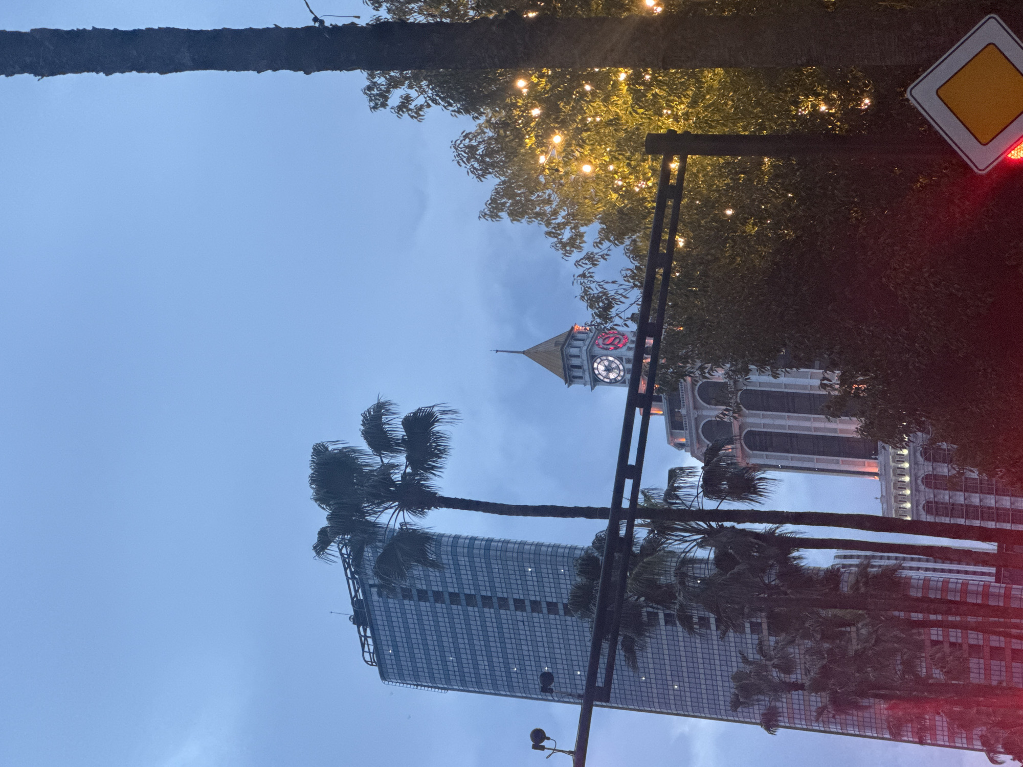 The image captures a cityscape featuring a mix of architectural styles. In the foreground, there are palm trees swaying in the wind. Behind the trees, two distinct buildings rise. One is a modern skyscraper with a glass facade, while the other is a more classical structure with a clock tower displaying the logo of the Sheraton hotel. Street lights and a yellow diamond-shaped traffic sign are visible. The sky is overcast, suggesting either early morning or evening. The image is taken from a low angle, looking upwards. The location is likely in Batumi, Georgia.