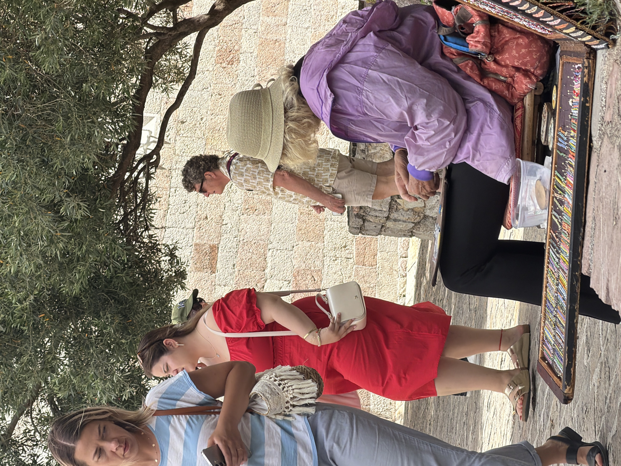 The image shows a street scene in Budva, Montenegro, with several people and objects. In the foreground, a person wearing a purple jacket and a straw hat is seated on a bench, with their hands engaged in an activity, possibly crafting or selling jewelry. In front of them is a display of colorful bracelets and other small items. To the left, a woman in a blue and white striped shirt and light-colored pants is holding a smartphone. Next to her, a woman in a bright red dress is walking, carrying a small white purse. Behind them, other individuals are visible in the background, including a man in a patterned shirt and shorts, and a person wearing a green cap. The environment appears to be an outdoor public space with a stone wall and a tree providing shade. The ground is paved with cobblestones.