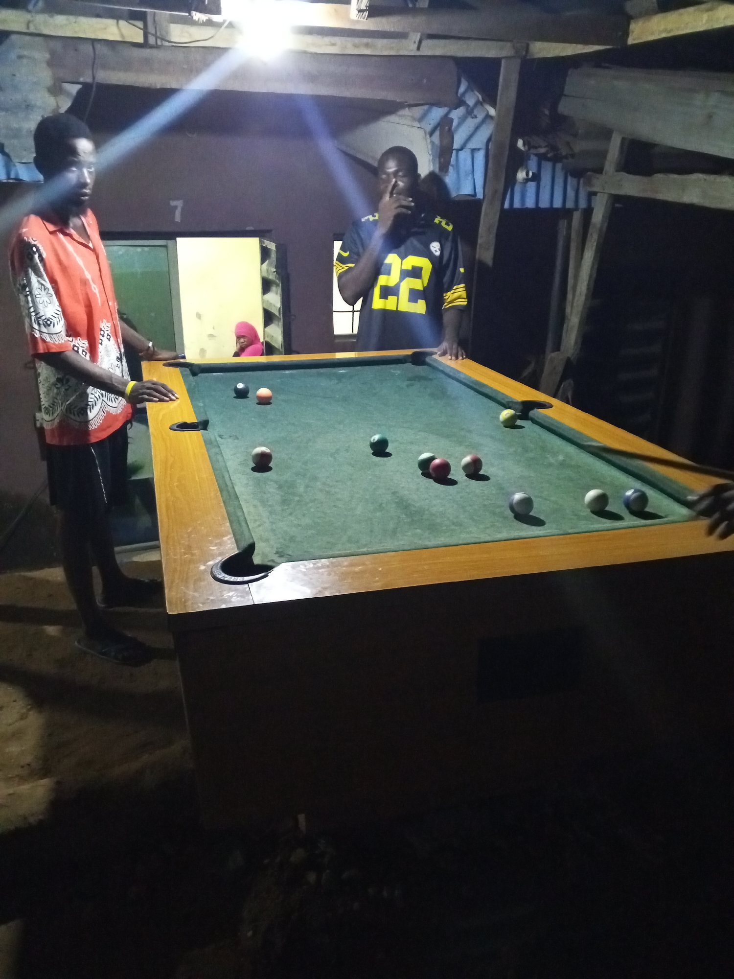 The image shows two men standing around a billiard table inside a dimly lit room, likely in Lokoja, Nigeria. One man, wearing an orange shirt with white patterns, stands to the left of the table. Another man, wearing a yellow and black sports jersey with the number 22, stands on the opposite side of the table, holding his hand to his mouth. The billiard table has a green felt surface with multiple billiard balls scattered across it. The room appears rudimentary, with exposed wooden beams and walls. A woman in a pink headscarf is partially visible through a doorway in the background. The scene is lit by an overhead light that shines brightly on the billiard table and the people around it.