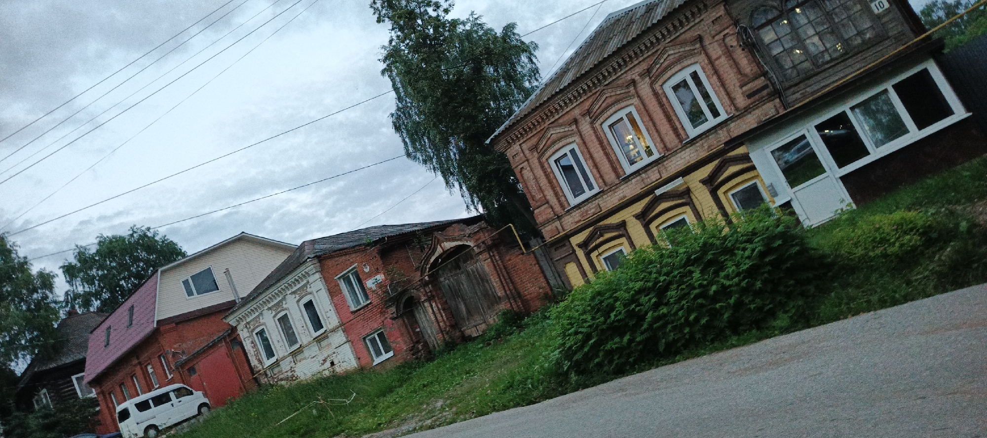 The image shows a street scene in Votkinsk, Russia, with several buildings of varying architectural styles. In the foreground, there is a gray asphalt road. To the left, a white minivan is parked in front of a red and white two-story house with a red roof. Further down the street, there is an older, weathered brick building with white trim and arched windows. Adjacent to this building is a two-story brick house painted in a reddish-brown hue with yellow decorative accents and white-trimmed windows. A dense green hedge runs along the front of this house. The background features a large, leafy tree and a cloudy sky with visible power lines. The overall impression is of a residential area in a Russian town.