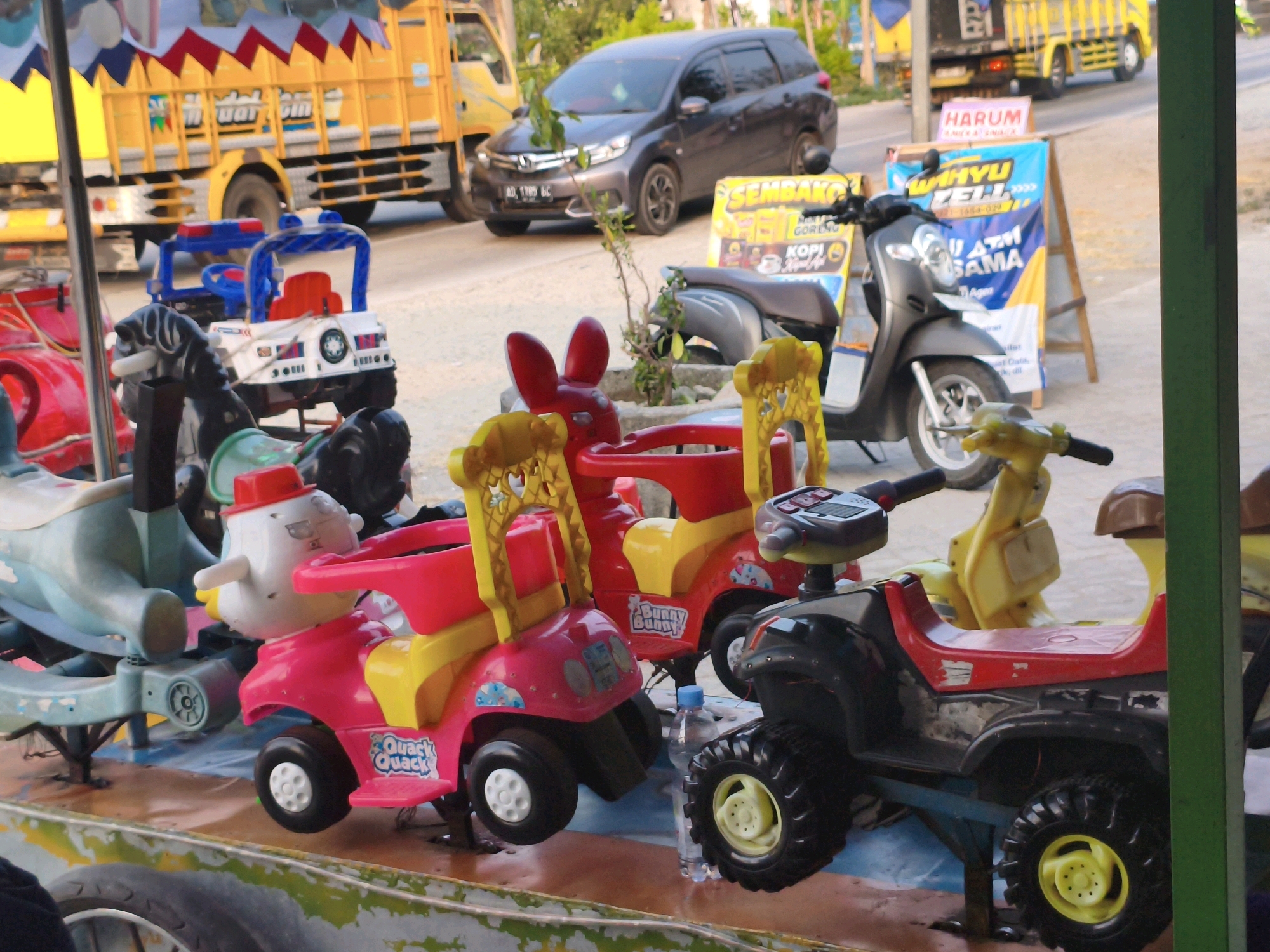The image shows a scene in Watubonang, Indonesia, featuring several colorful children's amusement rides, likely at a local event or park. In the foreground, a pink and yellow ride shaped like a duck with a red hat is prominently displayed. To its left, a blue and white police-themed ride is partially visible. Behind these, a dark gray car is parked on a street. Further in the background, a yellow truck and another vehicle are also present. The overall setting appears to be outdoors, with a mix of paved surfaces and natural elements.