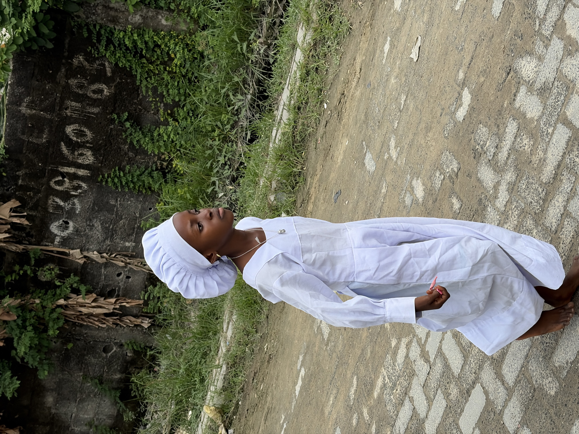 A young girl wearing a white cap and a white robe walks on a dirt path in Igboefon, Nigeria. The girl is positioned in the left half of the frame, facing to the right. Behind her, a weathered concrete wall is visible with graffiti and some dried plant material hanging from it. Lush green vegetation grows along the base of the wall and further back. The ground in the foreground consists of a dirt path interspersed with cobblestone-like pavers. The lighting suggests an overcast day or a shaded area.