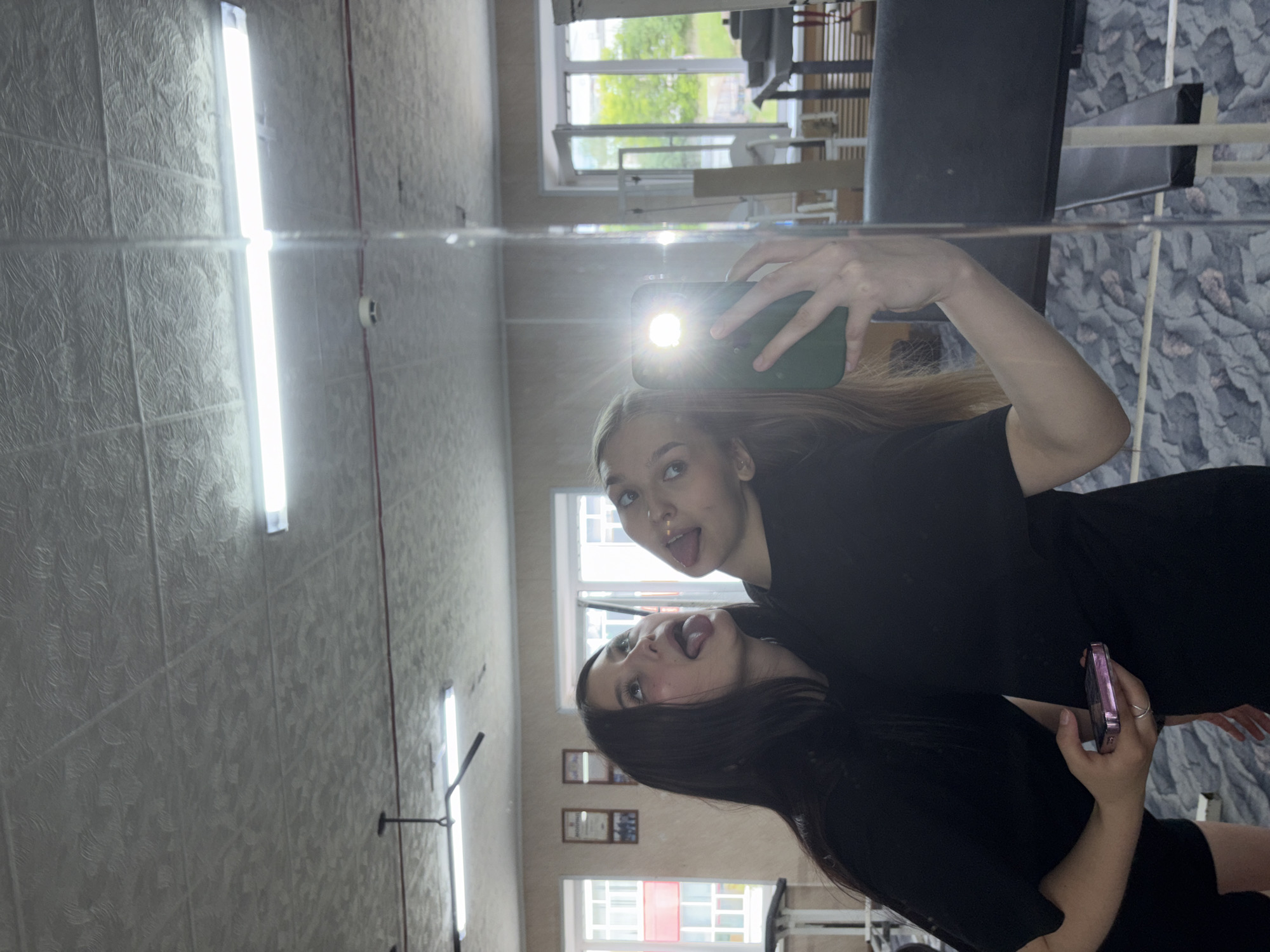The image shows two young women taking a selfie in a mirrored room, likely a gym, as evidenced by the workout equipment visible in the reflection. The woman on the left, with dark hair, leans towards the woman on the right, with lighter hair, sticking her tongue out. The woman on the right is holding a green-cased smartphone, capturing the reflection. The background shows fluorescent lighting fixtures on a textured ceiling and windows with a view of trees and buildings, suggesting an urban environment. The floor has a patterned design. The overall atmosphere is playful and casual. The location is the city of Chaikovsky, Russia.