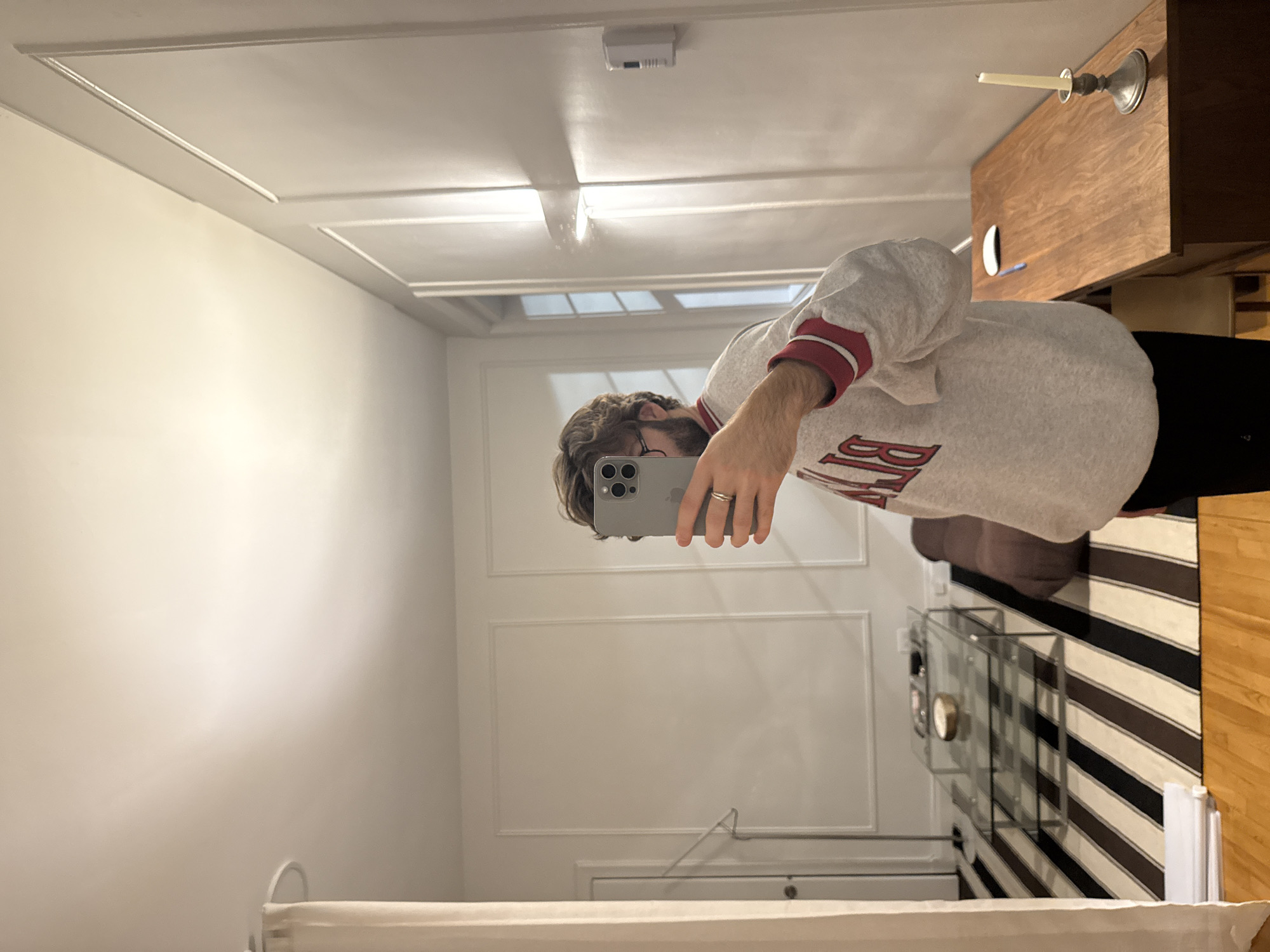 A man is taking a selfie in a mirror. The reflection shows a room with white walls, a bed in the foreground, and a glass coffee table on a striped rug in the background. A wooden cabinet with a candle holder sits in the upper right corner. The location appears to be an apartment interior, potentially in Montreal based on architectural styles common in that city.