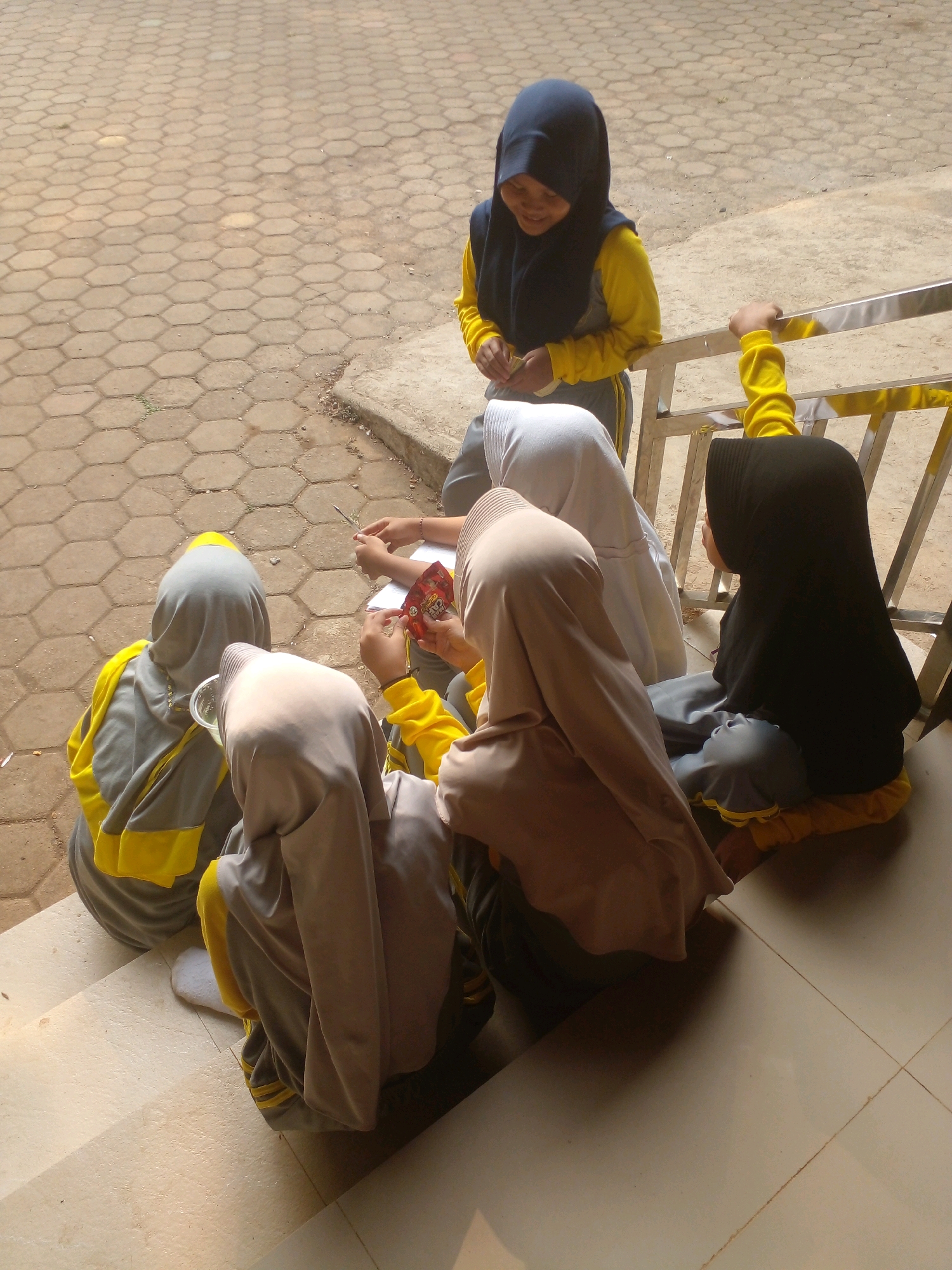 In Banyuputih, Indonesia, six young girls, all wearing hijabs and school uniforms with yellow and gray accents, are gathered on outdoor concrete steps. One girl in a navy hijab and yellow shirt is standing, looking down and holding a small object. The other five girls are seated on the steps. Three of them are facing away from the camera, appearing to be engaged in an activity, with one holding a red packet and another holding what seems to be a clear plastic cup. Another girl in a white hijab is partially visible, and a girl in a black hijab is seated on the right, looking to the side. The environment suggests a sunny day, with shadows cast on the hexagonal-patterned ground and the steps. A metal railing is present to the right of the standing girl.