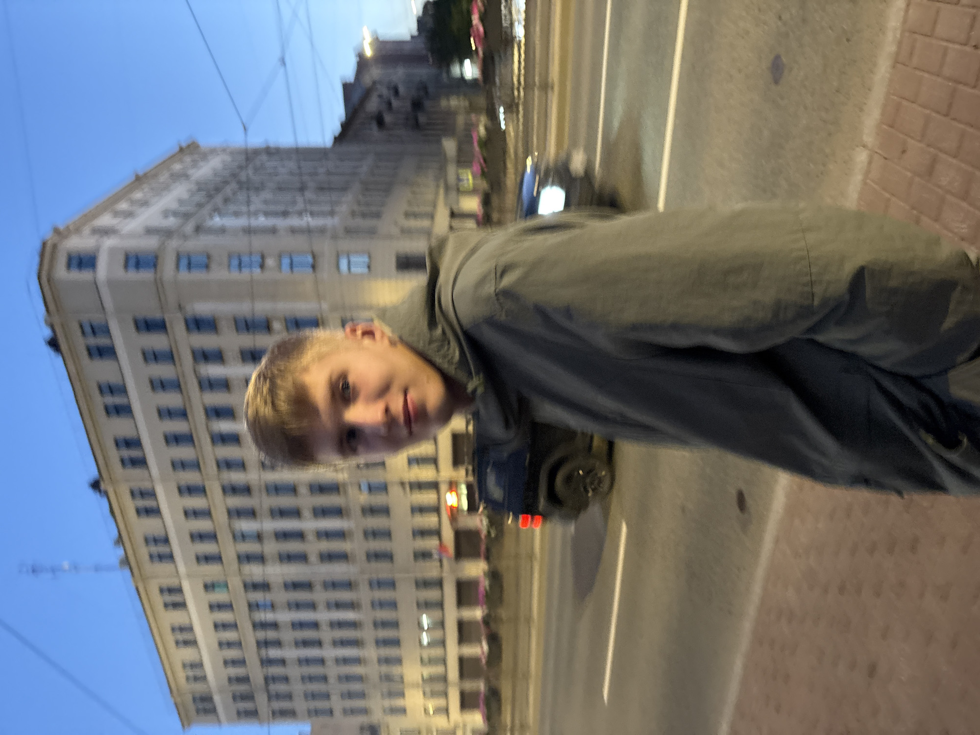 A medium shot captures a young Caucasian male with light blonde hair and fair skin in the foreground, looking towards the camera. He is wearing a dark green hooded jacket. The background features a multi-story building with numerous windows and balconies, characteristic of St. Petersburg's historic architecture, bathed in the twilight glow. The building appears to be on a wide avenue, with a road and sidewalk visible in front of it. Traffic, including a dark-colored SUV and other vehicles, is present on the road, with some lights illuminated. Planters with flowers are visible along the sidewalk in front of the building. The sky is a deep blue, suggesting dusk or early evening.