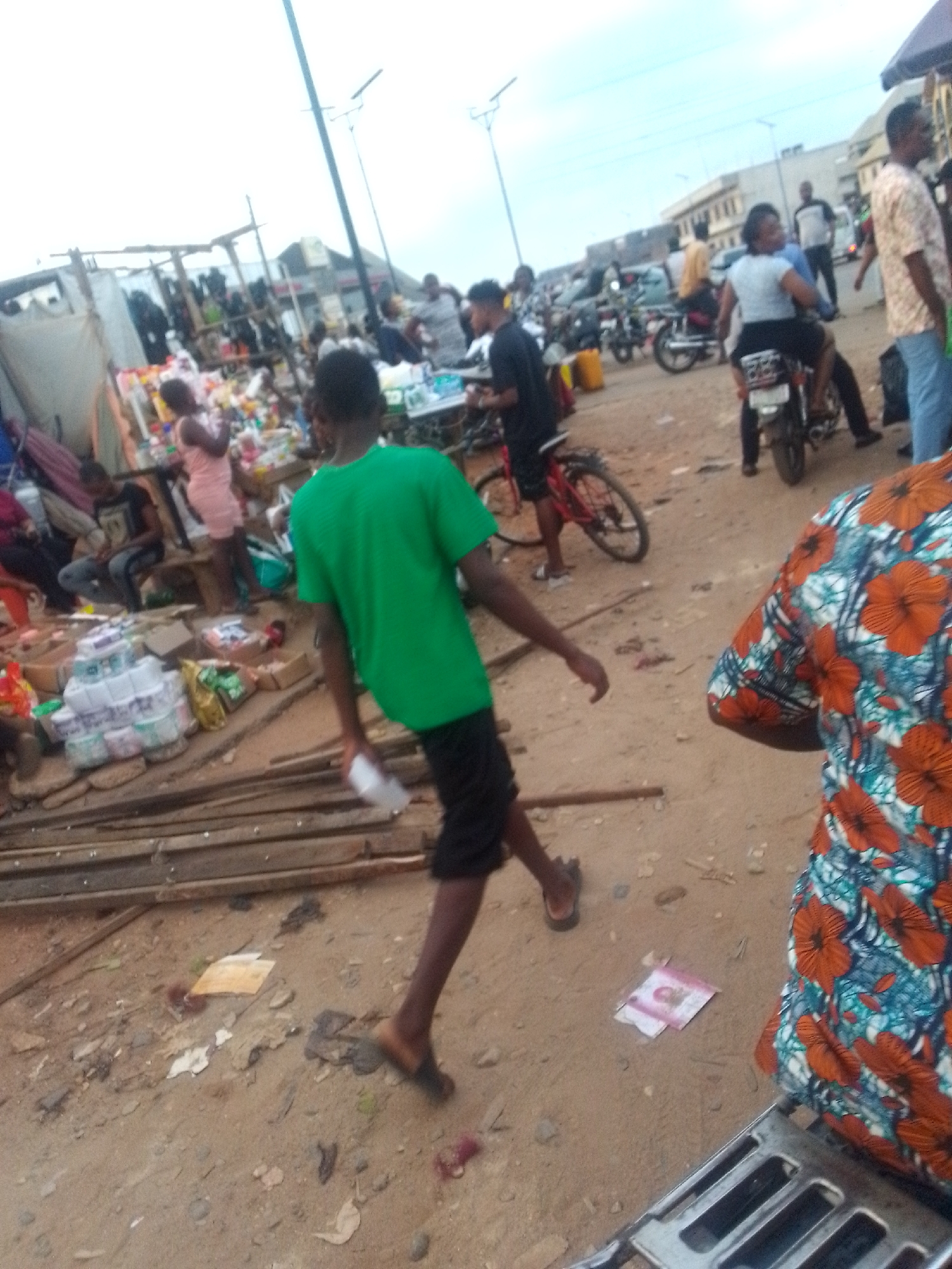 The image shows a bustling street scene in what appears to be a marketplace. A person in a green shirt and black shorts is walking away from the viewer. Several people are gathered around stalls selling various goods. A woman is riding on the back of a motorcycle. Other people are walking or standing nearby. There are bikes, motorcycles, and cars parked along the street. The ground is dirt or sand, and there are some scattered debris. The sky is overcast.
