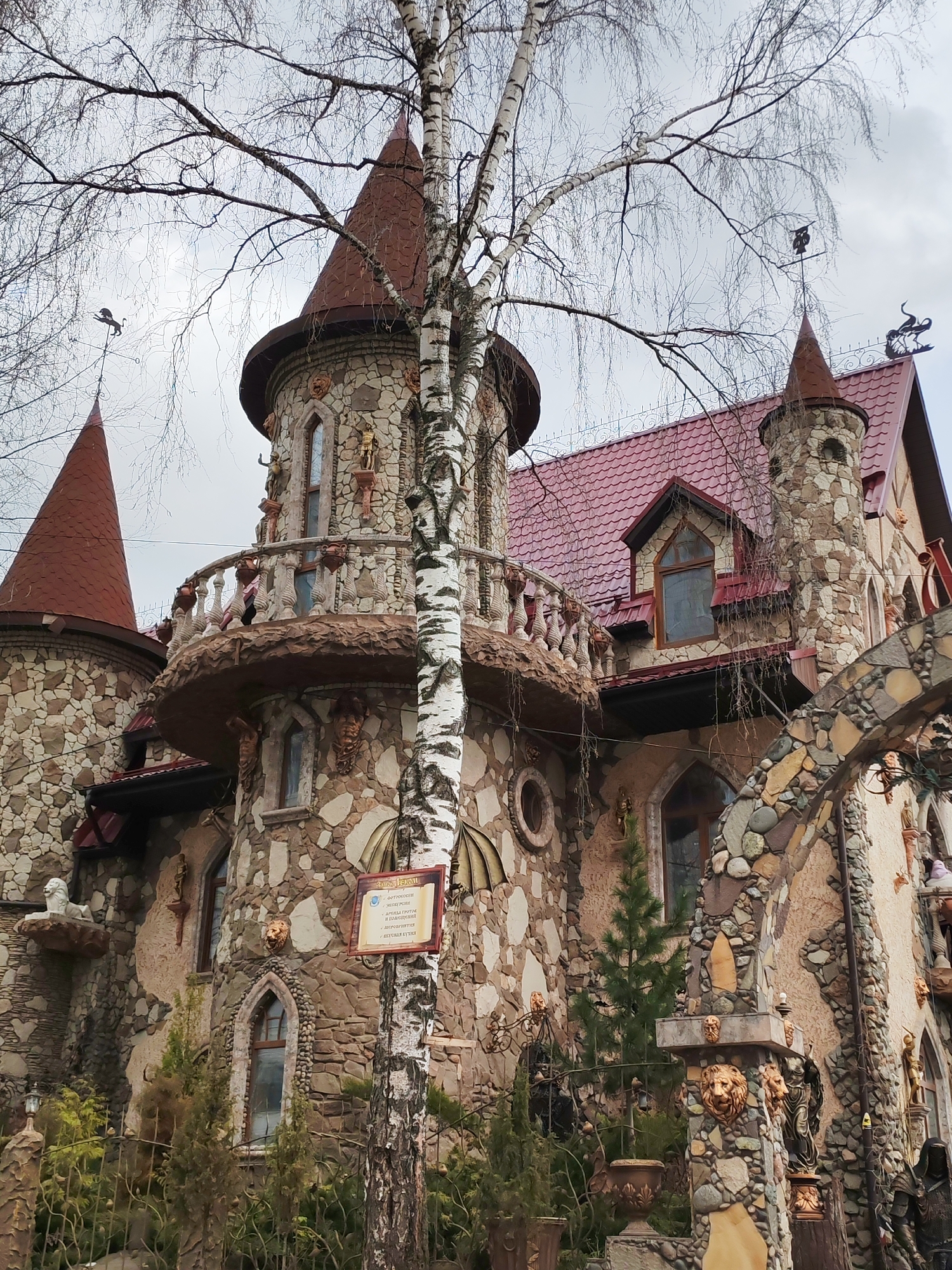 The image captures a whimsical, castle-like building in Izhevsk, Russia, with multiple turrets and red conical roofs, set against an overcast sky. A tall, bare birch tree stands prominently in the foreground, its leafless branches partially framing the structure.
The building's facade is intricately constructed from diverse natural stones, creating a textured, mosaic-like appearance. Architectural details include arched windows, a prominent rounded balcony with balusters adorned with decorative figures, and various small sculptures of human figures and animals integrated into the stonework. Weather vanes, including one shaped like a dragon or griffin on a left turret and another resembling an owl on a right turret, crown some of the rooftops. A larger, more elaborate weather vane depicting a mythical creature is visible on the main roofline on the far right.
At ground level, the area around the building features sparse greenery, including small evergreen shrubs, and ornate fences embellished with lion head decorations. An arched gateway, also made of varied stones, provides access. Sculptures of lion heads and other figures are visible on pillars and walls.
A wooden sign attached to the birch tree provides context. Written in Russian, it is titled "Замок Веном" (Venom Castle) and lists services: "Фотосессии" (Photoshoots), "Декорации" (Decorations), "Съемки клипов и объявлений" (Filming of clips and announcements), "Мероприятия" (Events), and "Кастинг студии" (Casting studio). This indicates the building functions as a commercial or creative venue.
The time of day is daytime, under a cloudy sky, suggesting a muted light. The bare branches of the birch tree indicate either late autumn, winter, or early spring. No people or specific activities are visible, emphasizing the unique architecture of the building.