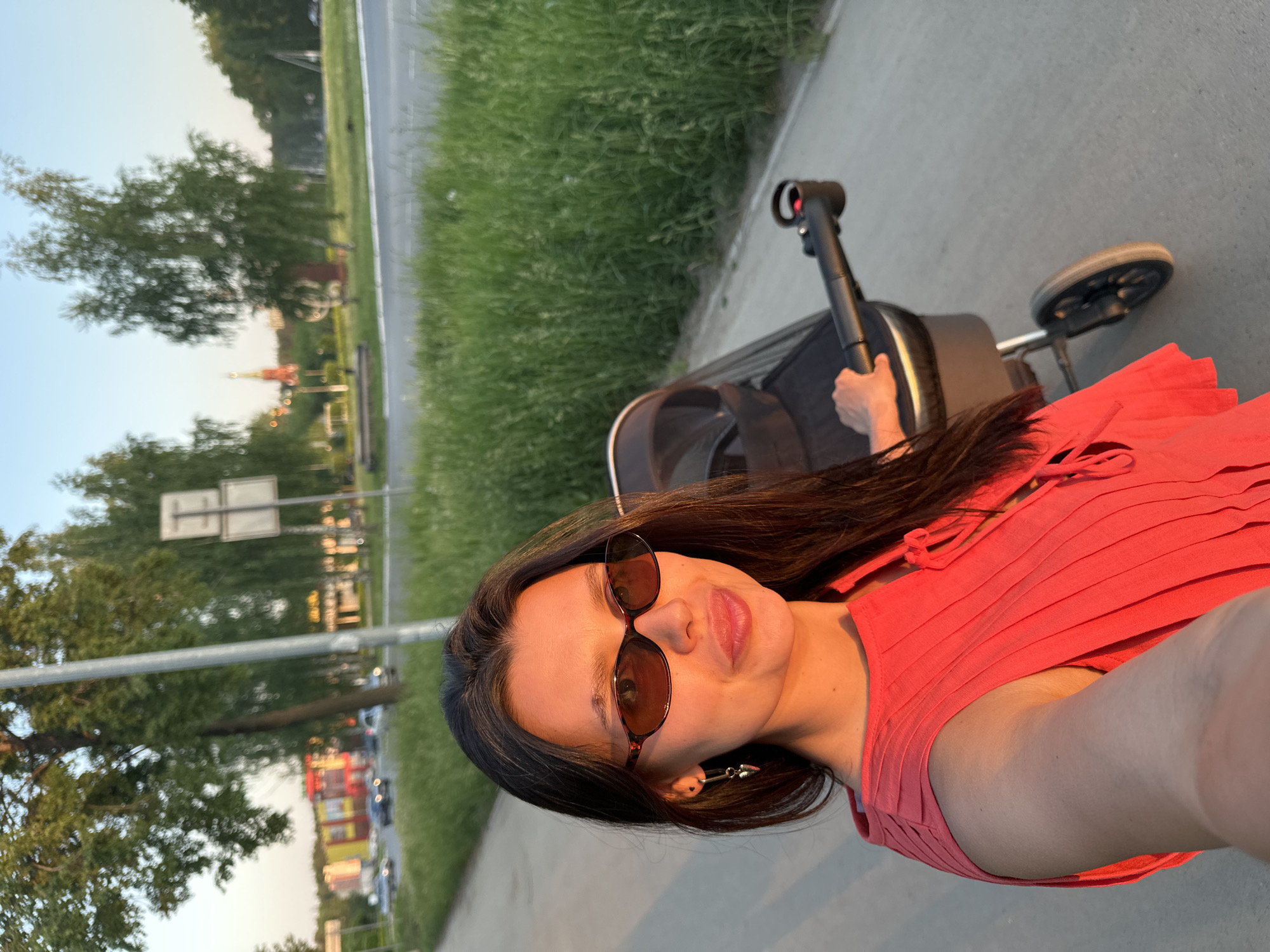A woman is taking a selfie outdoors. She is wearing sunglasses and a red top. She is standing next to a baby stroller on a paved sidewalk. Beyond the sidewalk is a grassy area and a road. In the distance, buildings and trees are visible. The woman is likely in Chaikovsky, Russia, based on the presence of a building with a spire that resembles the architecture of the city. The photo is taken during the daytime.