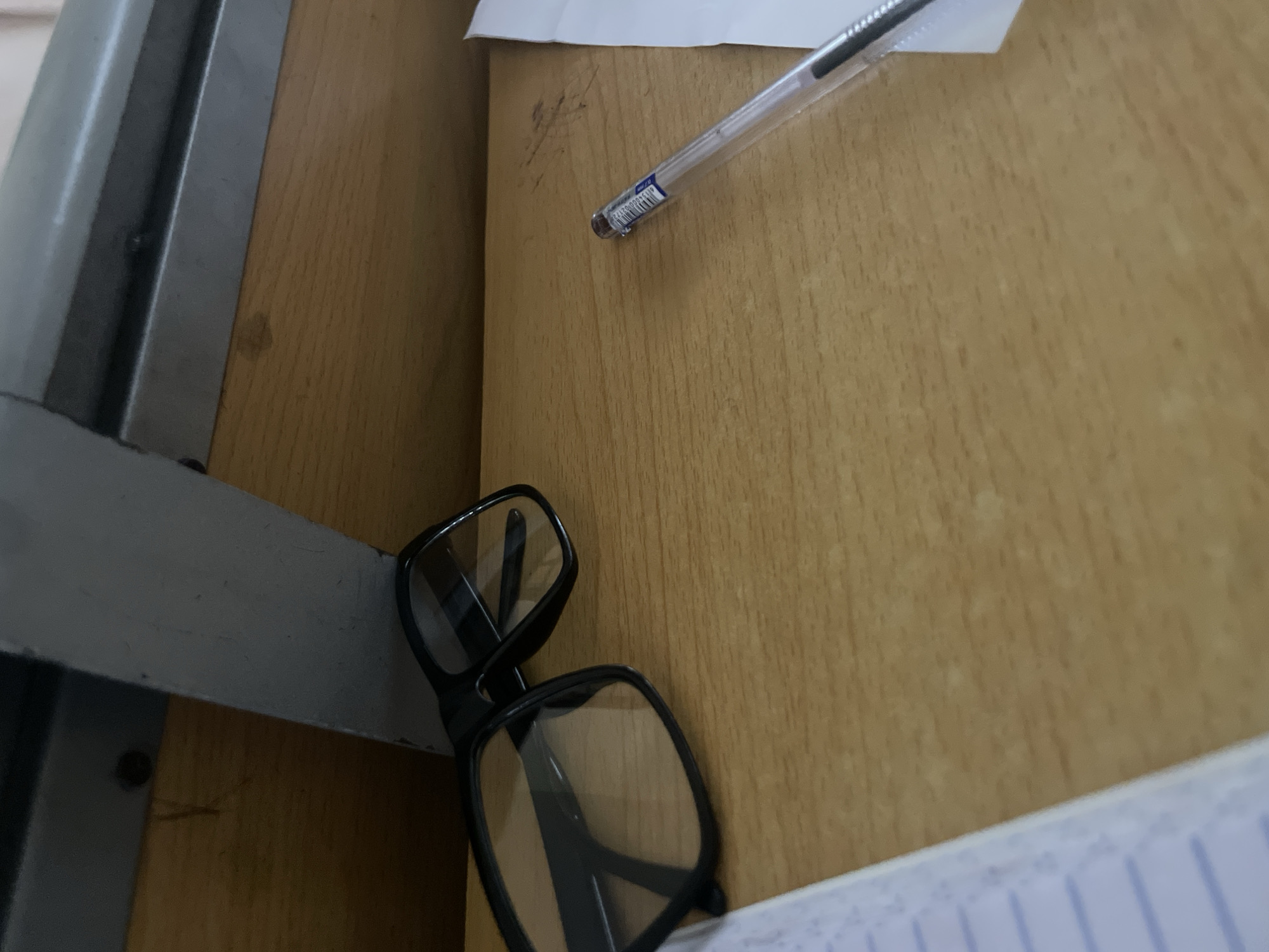 The image shows a close-up, eye-level shot of a wooden desk surface. On the left side of the desk, a pair of black-framed glasses is laid down. To the right of the glasses, a clear plastic pen with a blue ink cartridge is positioned diagonally. In the background, a metallic structure is visible, suggesting the desk is part of a larger piece of furniture, possibly a school desk with an attached metal frame. A corner of a notebook with ruled lines is visible on the far left edge of the frame. The lighting suggests an indoor environment, likely during the day. The scene is static and appears to depict a workspace. There is no information about the location in Ihiagwa, Nigeria.